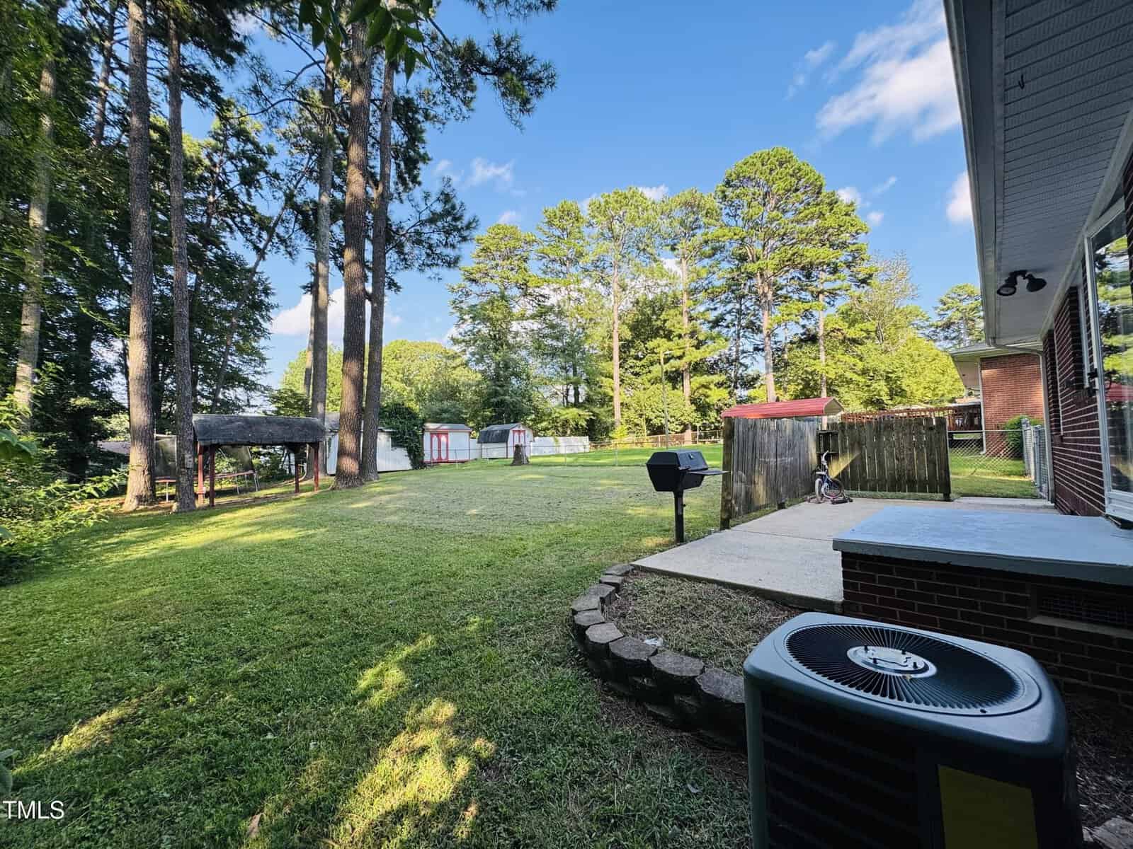 A grassy backyard with tall trees, sheds, a small playground, a grill on a concrete patio, and an air conditioning unit beside a brick house under a blue sky.