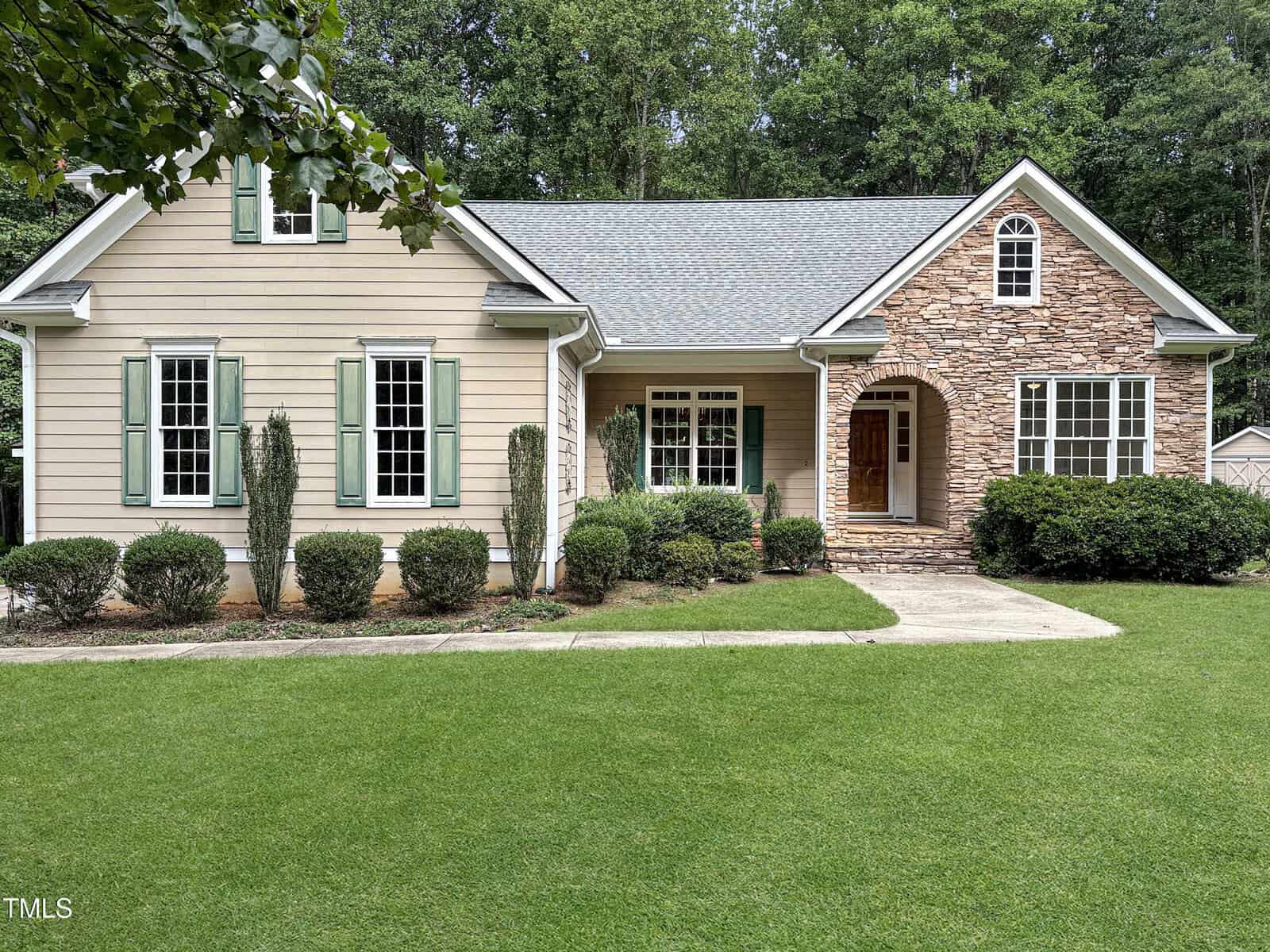 Single-story house with beige siding, green shutters, and a front entrance accented by stonework. The yard is well-maintained with shrubs and a lush green lawn, bordered by trees in the background.