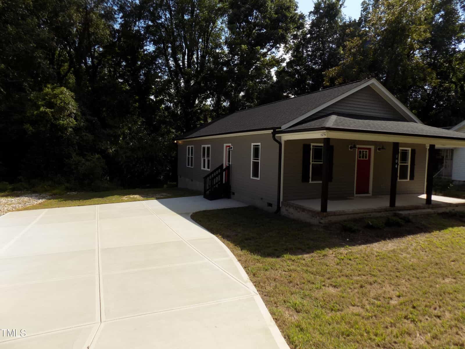 Single-story gray house with a covered front porch, black shutters, red door, and a ramp. There is a large concrete driveway and yard, with trees and woods in the background.