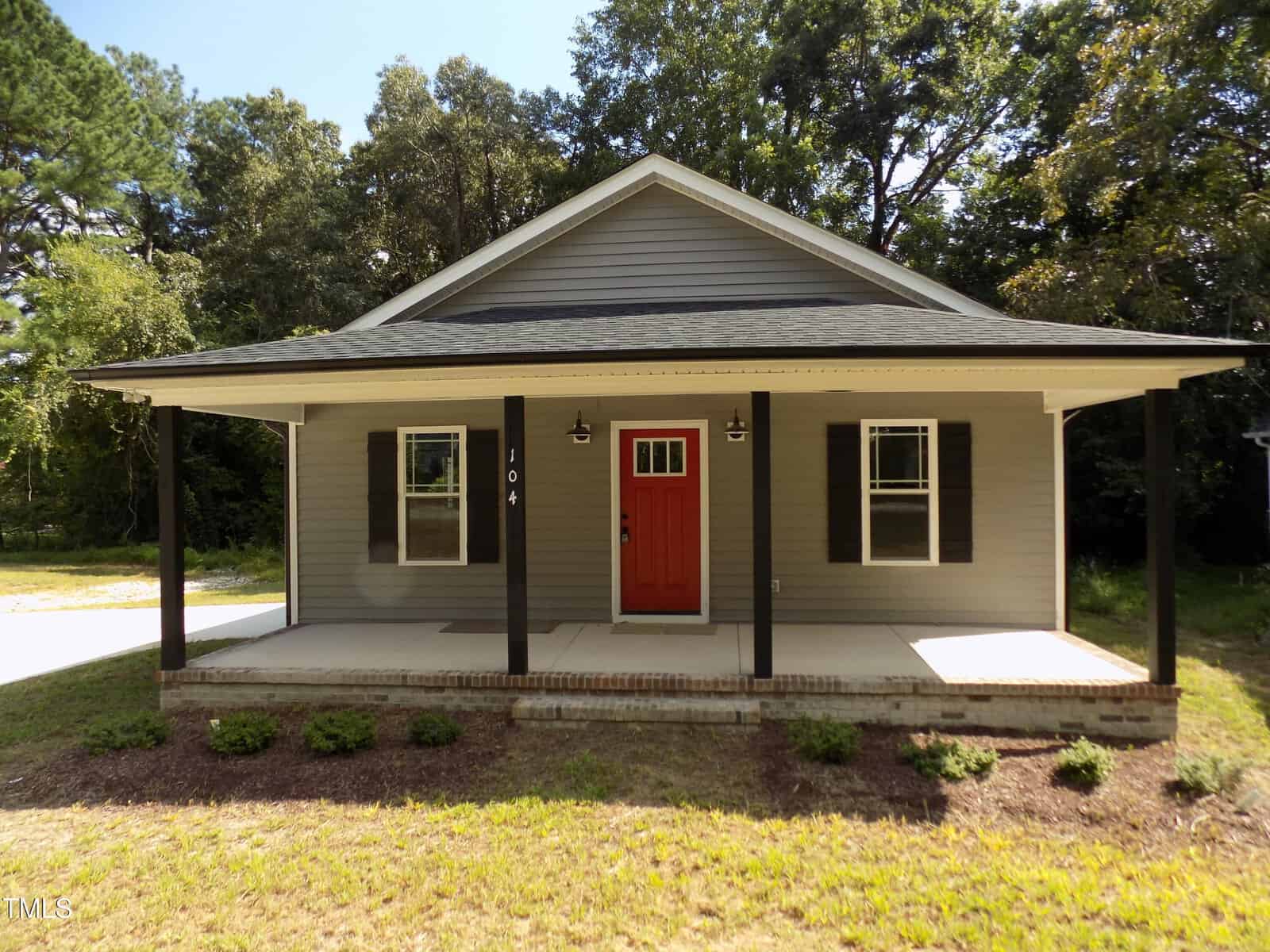 Single-story house with gray siding, black shutters, and a red front door located at 104 Halifax Road in Louisburg. The home features a covered front porch with four black columns and a small garden bed along the front. Trees and grass surround the property.