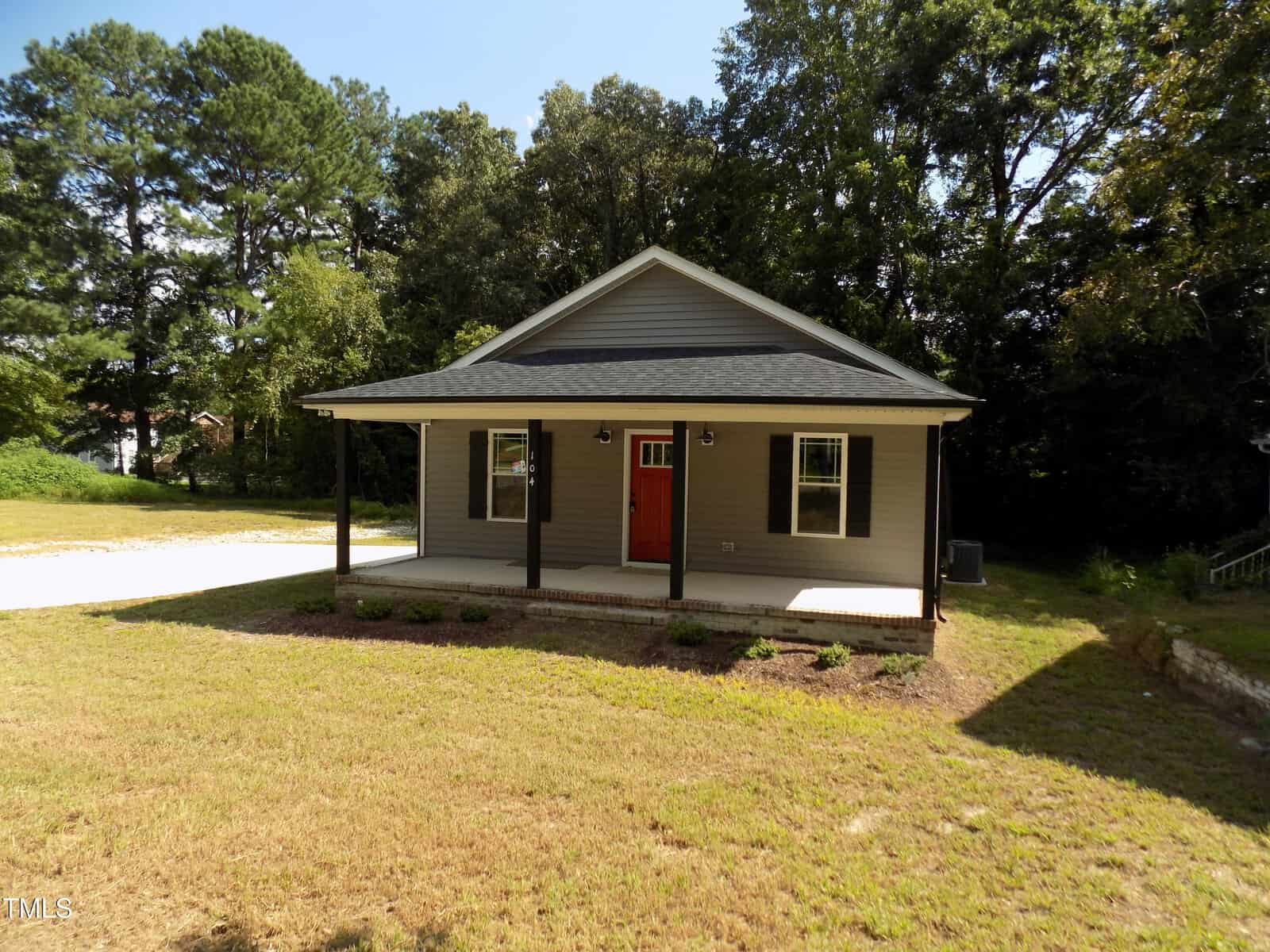 A single-story house with gray siding, black trim, a covered front porch, and a red door sits on a grassy lot with a concrete driveway and surrounded by tall trees.