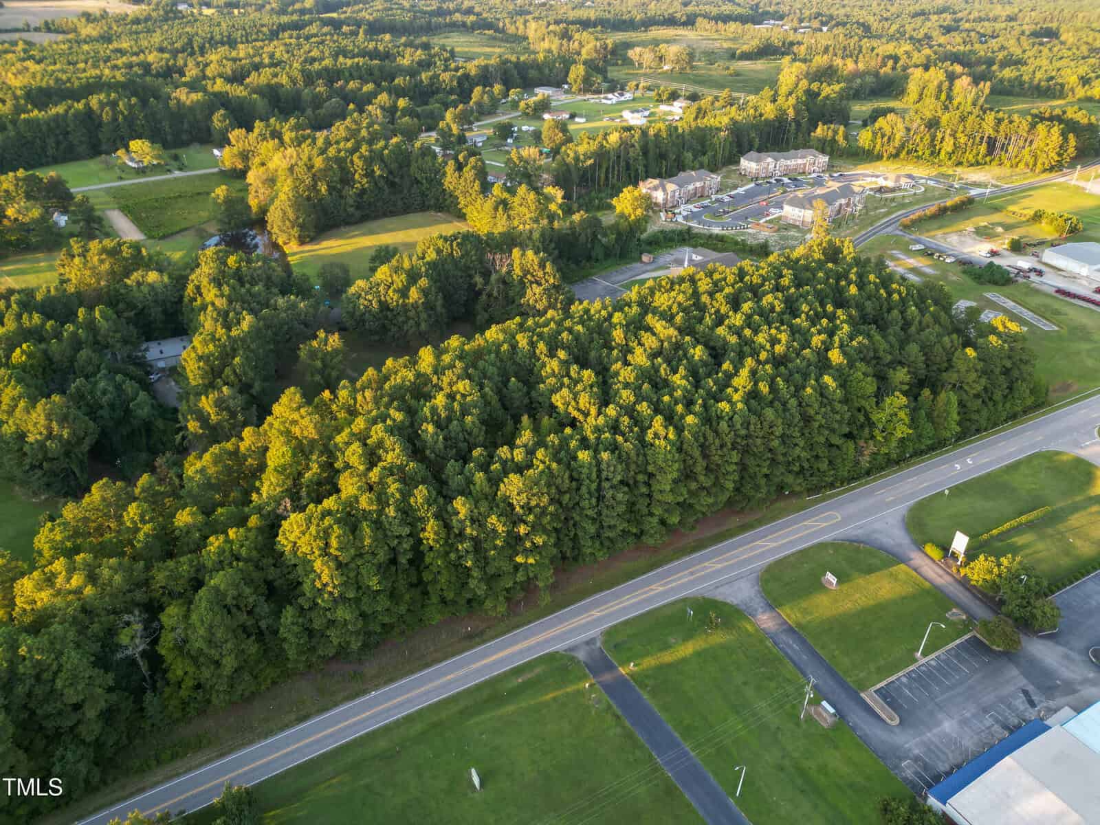Aerial view of a tree-covered area bordered by a paved road in the foreground, with grassy fields, scattered buildings, and a residential development in the background. Dense forest occupies most of the center of the image.