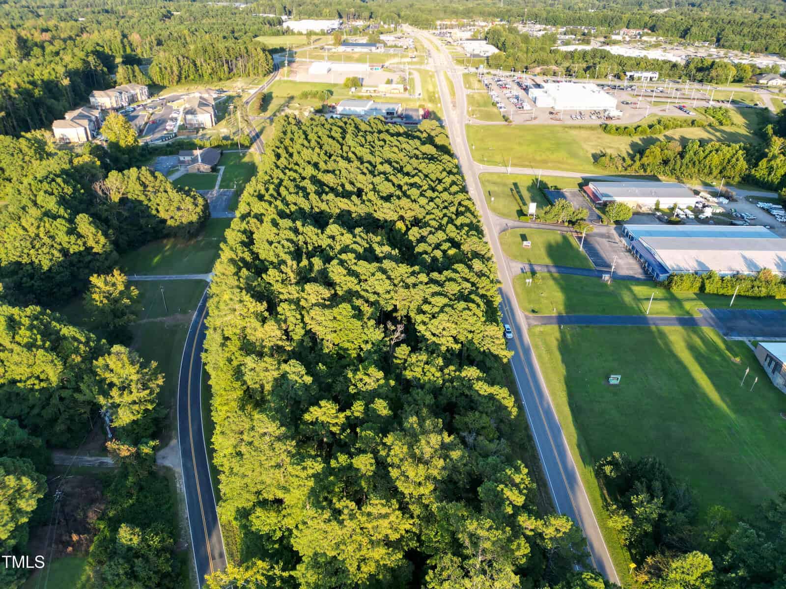Aerial view of a forested area bordered by roads near 01 158 Business Hwy, Henderson, with commercial and industrial buildings, parking lots, and open grassy fields surrounding the woods. The landscape stretches to the horizon under bright daylight.