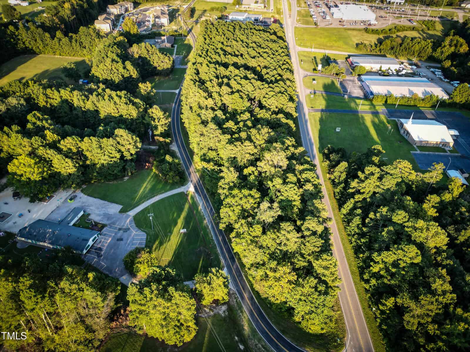 Aerial view of a winding road cutting through a dense, green forest area, with buildings, parking lots, and grassy fields visible on either side. Sunlight casts shadows from the trees across the landscape.