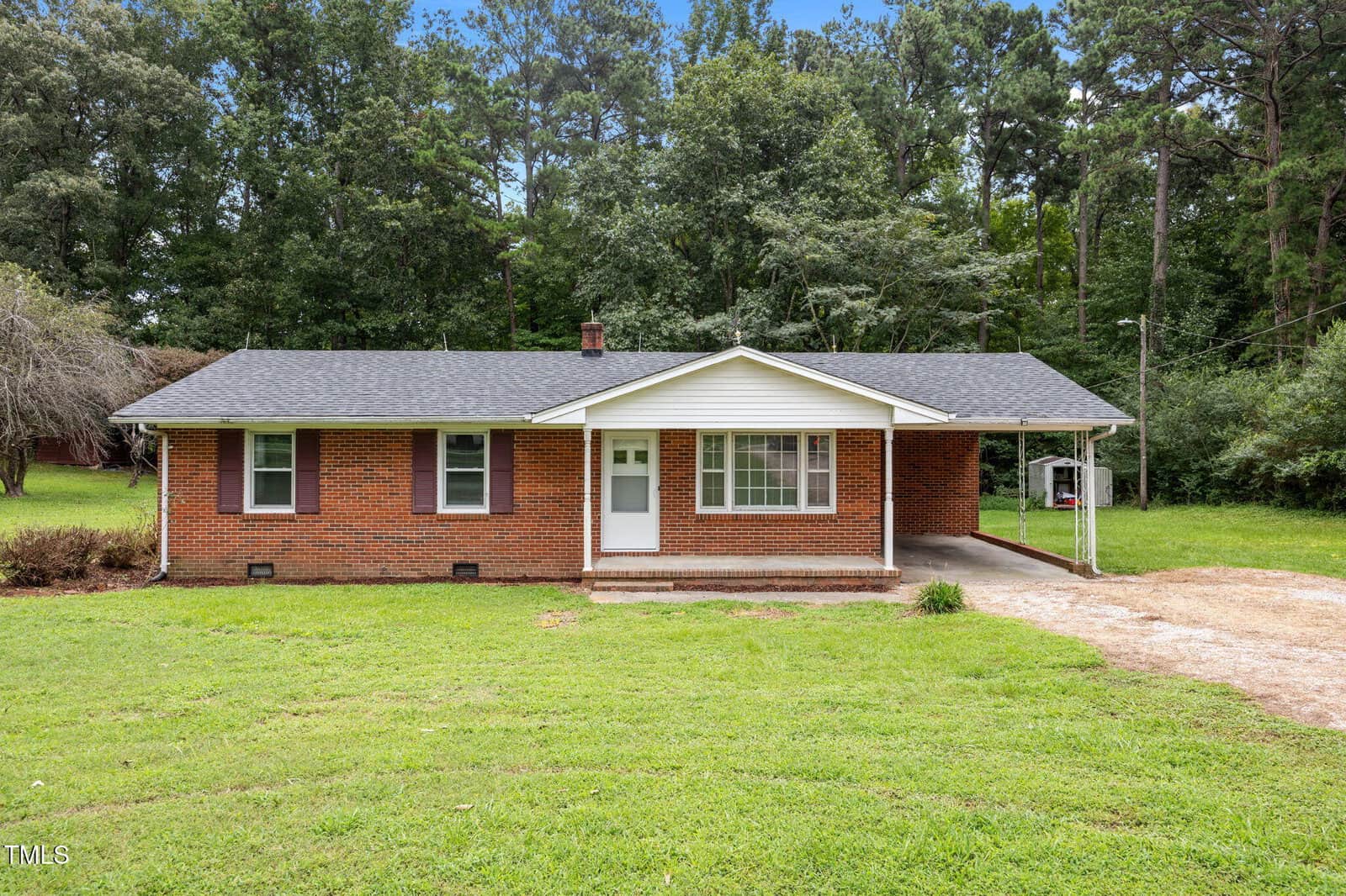 A single-story brick house at 1050 Rock Spring Church Road in Henderson features a dark shingled roof, white trim, and a covered carport on the right side. Set on a grassy lawn, it's surrounded by tall trees in the background.