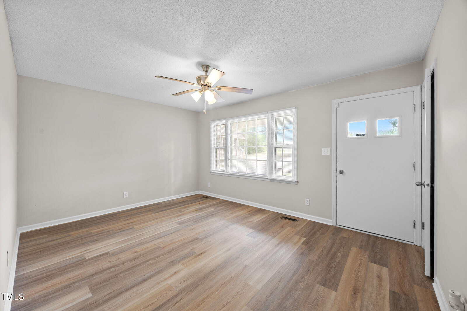 A room with light-colored walls, wood-look flooring, a ceiling fan with lights, a window, and a white front door with two square windows, viewed in daylight. The room appears empty and clean.