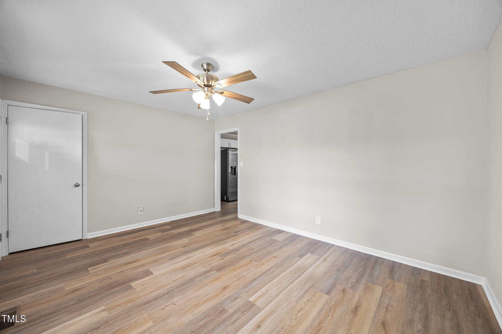 Empty room with light-colored laminate flooring, beige walls, a white ceiling, a ceiling fan with lights, a closed white door, and an open doorway leading to a kitchen with a visible refrigerator.