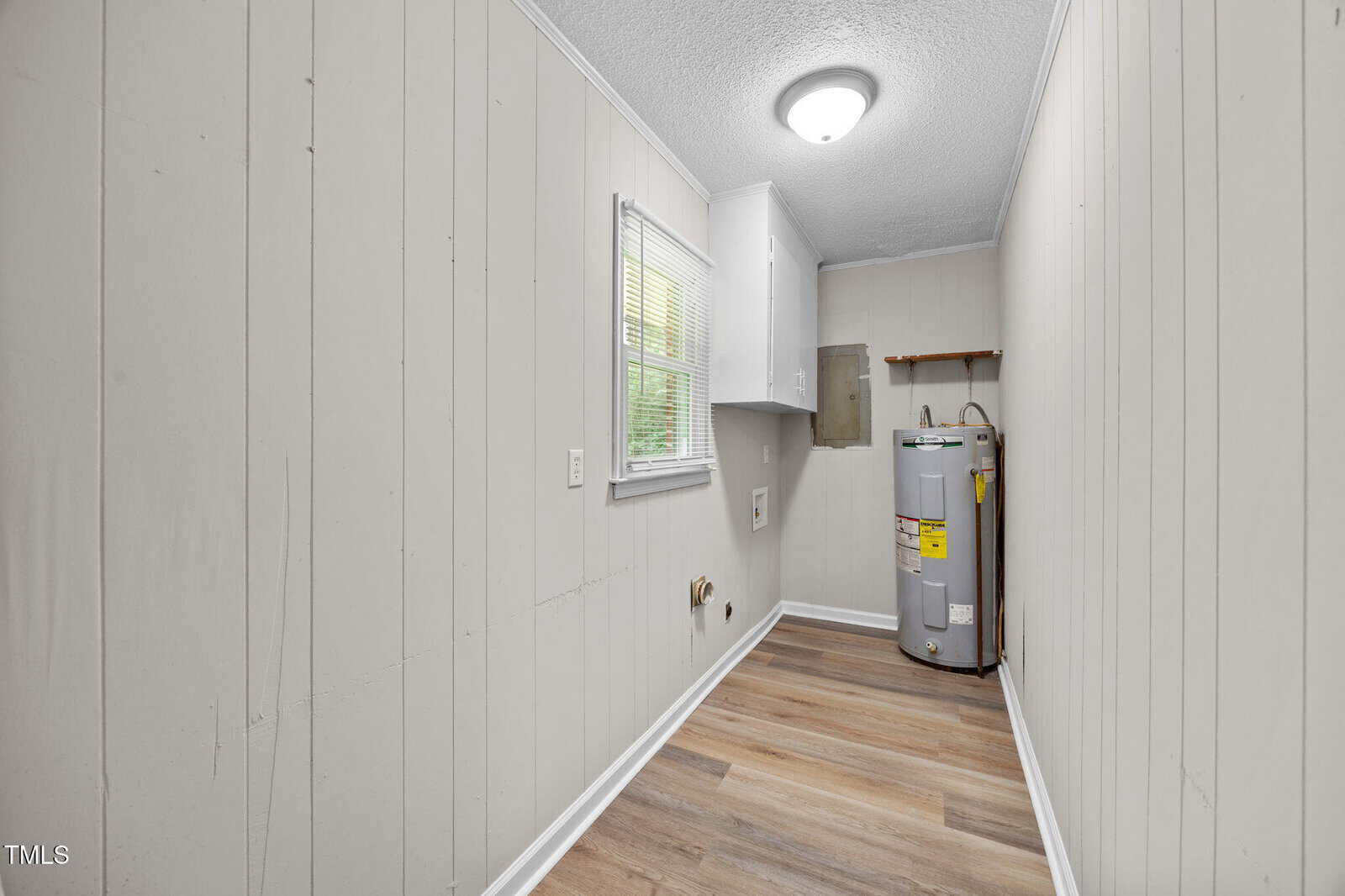 A narrow laundry room with light-colored wood panel walls, a window, a ceiling light, upper cabinets, hookups for washer and dryer, and a water heater on the right side near the back wall.