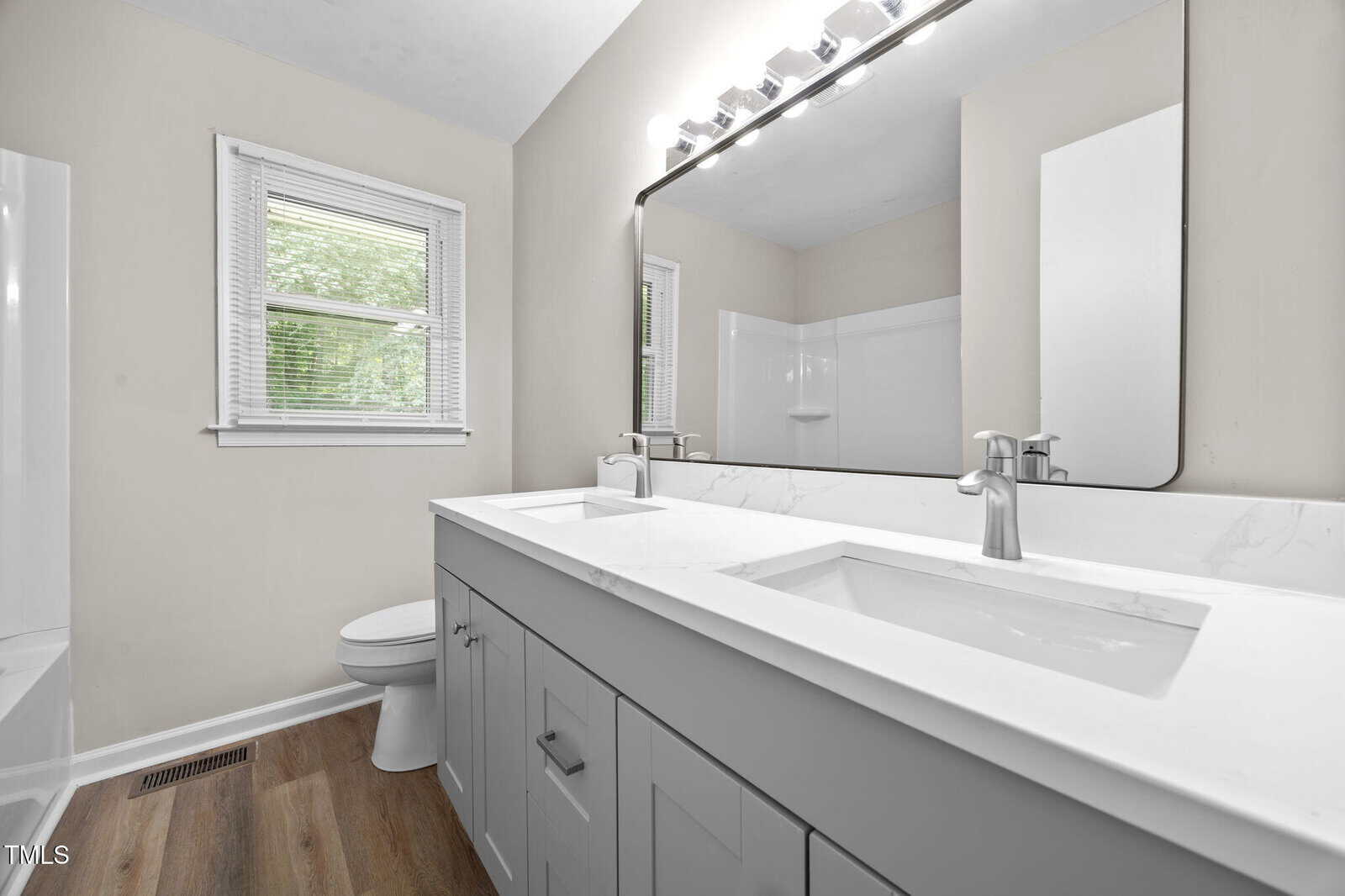 Modern bathroom with a double sink vanity, large wall mirror, white countertops, light fixtures, a toilet, a window with blinds, and wood-style flooring. Neutral wall color enhances the clean look.