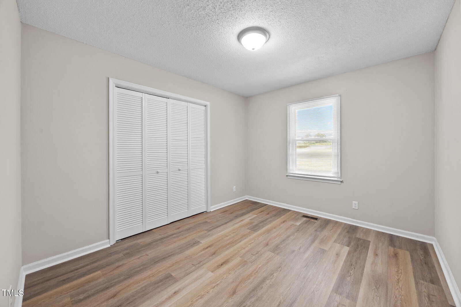 Empty room with light beige walls, a ceiling light fixture, a window with white trim, a closed white double-door closet, and wood-look laminate flooring.