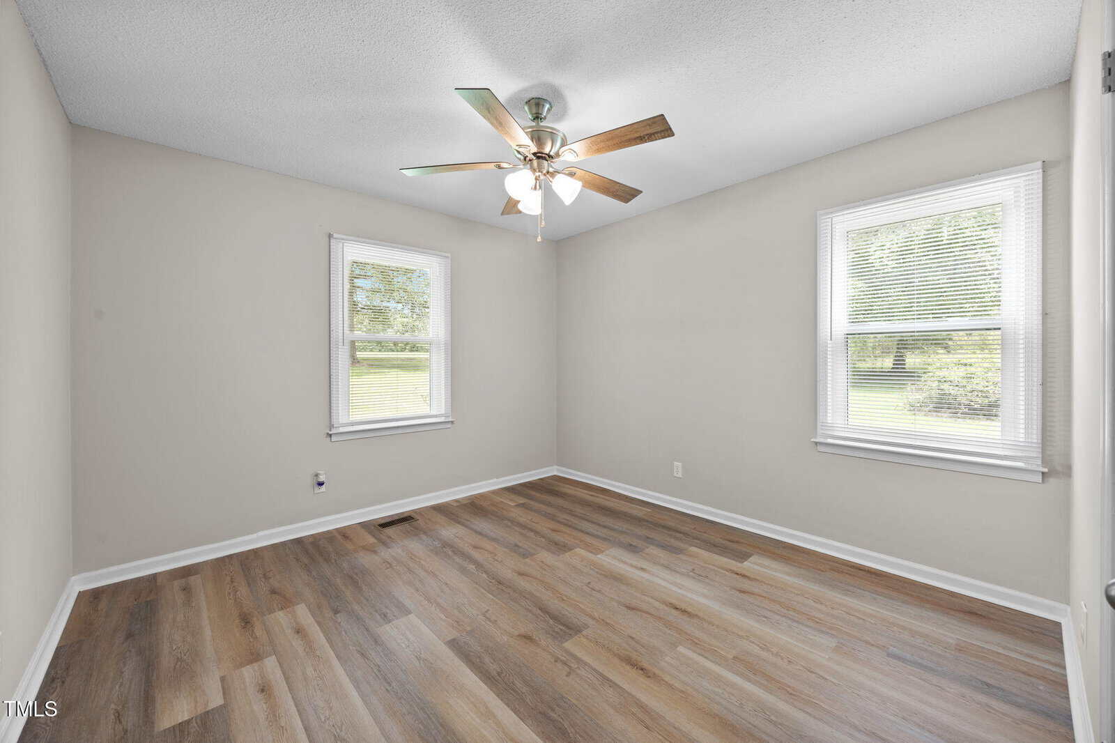 An empty room with light gray walls, two windows with blinds, a ceiling fan with lights, and wood-look vinyl flooring. Natural light enters through the windows, revealing a view of green grass and trees outside.