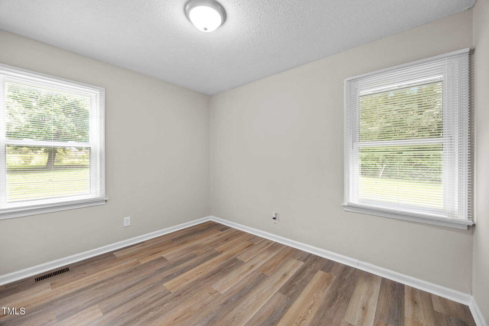 Empty room with light gray walls, two large windows with blinds, wood-look vinyl flooring, white ceiling, and a round ceiling light. Sunlight is coming in from both windows, and outside greenery is visible.