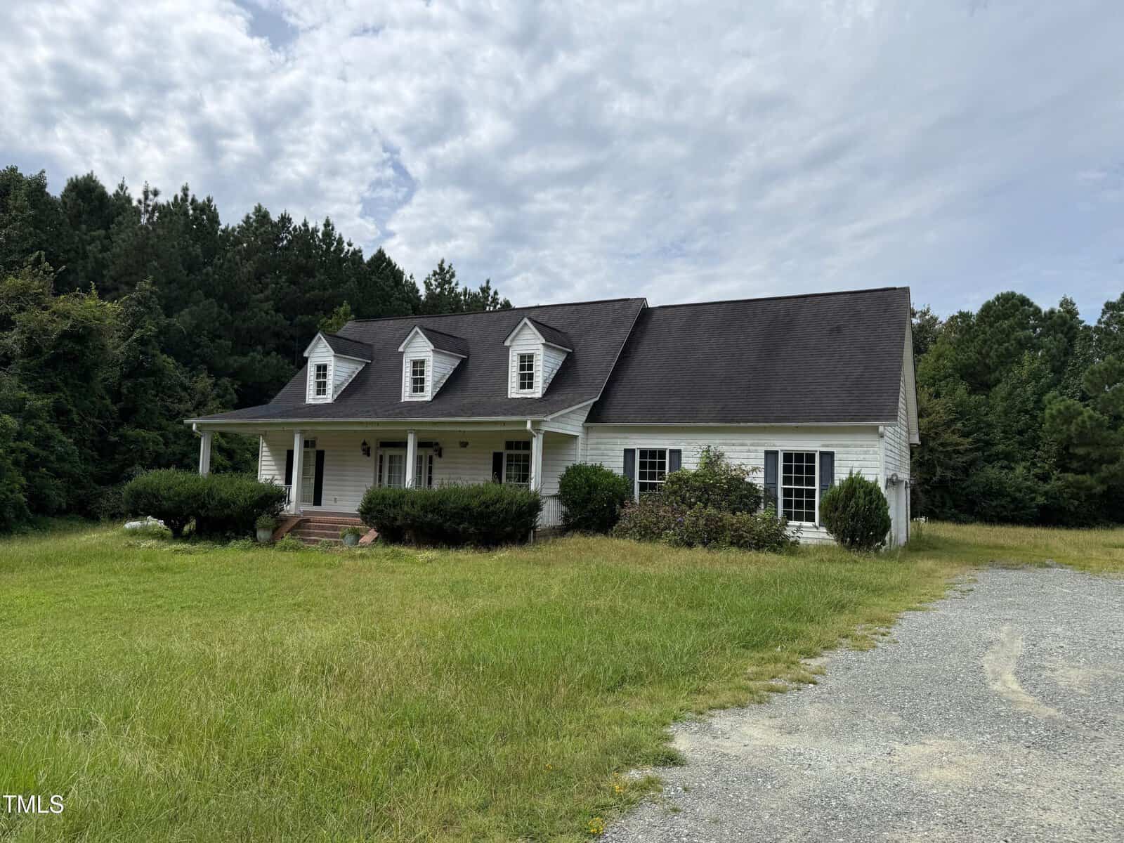 A white, two-story house with three dormer windows and a covered front porch sits on a grassy lot at 1115 Will Suitt Road in Creedmoor, nestled among trees beside a gravel driveway under a cloudy sky.