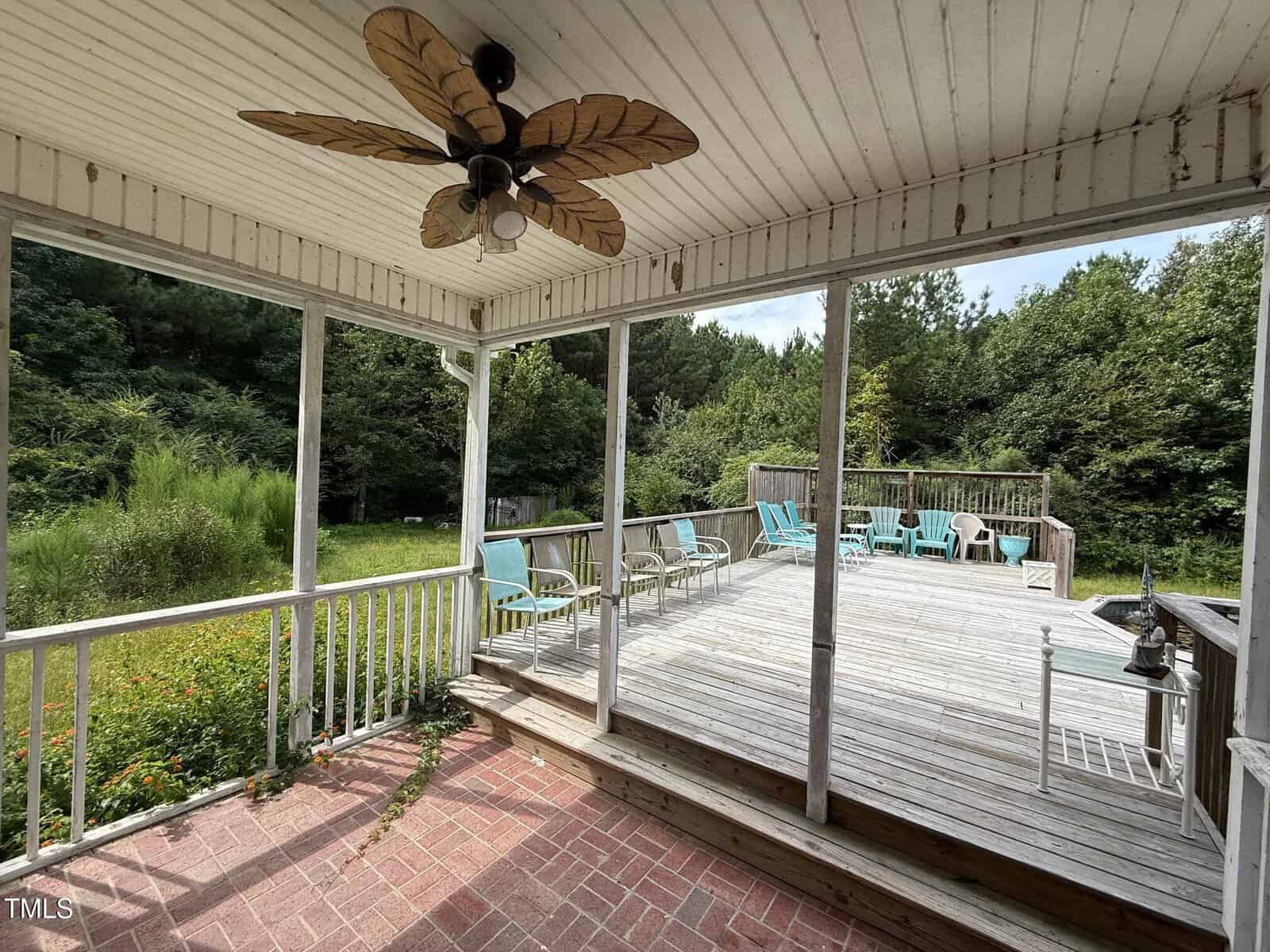 A covered porch with a ceiling fan opens to a large wooden deck with outdoor seating. The deck overlooks a grassy backyard bordered by dense trees and shrubs.