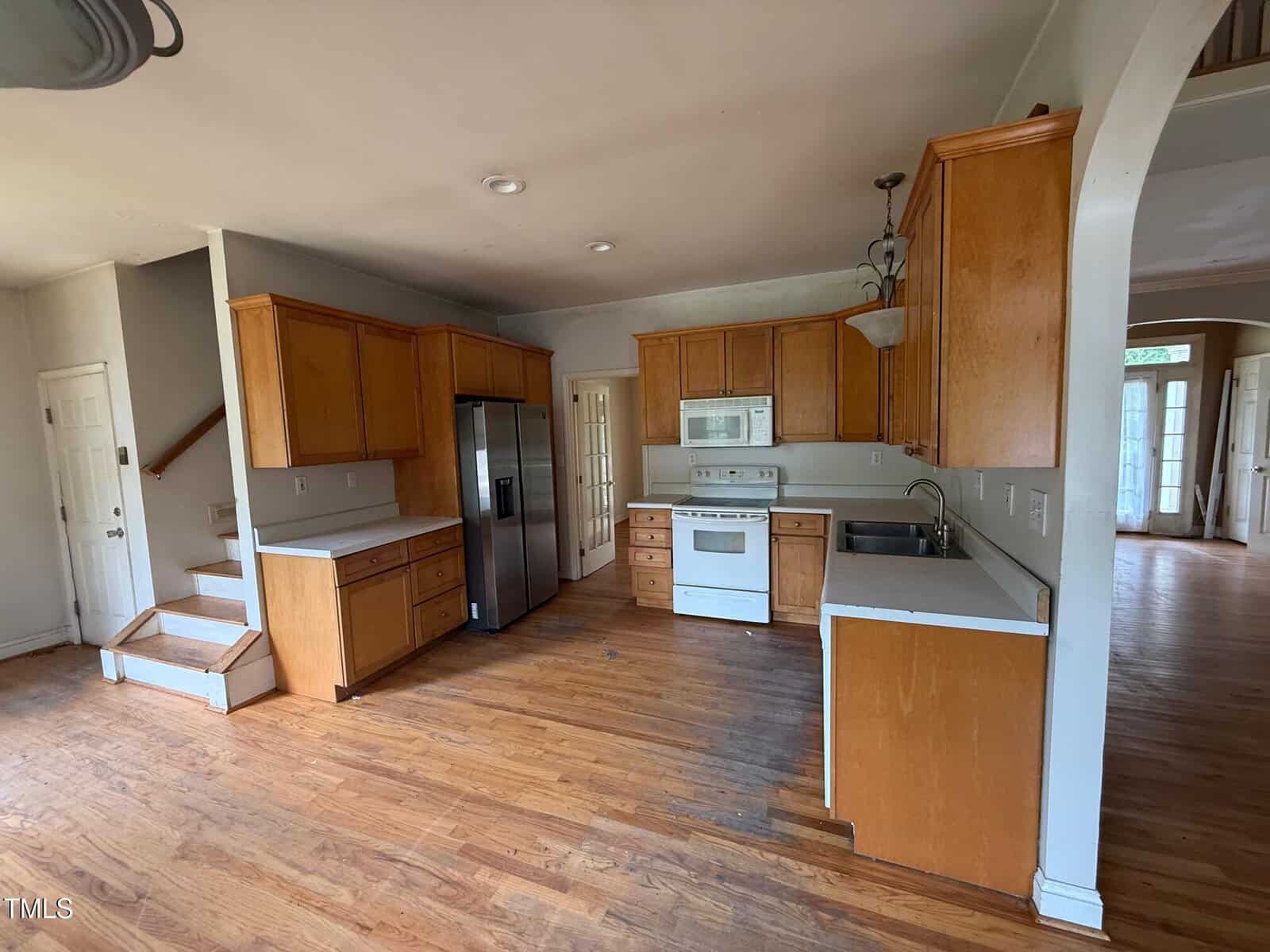 A kitchen with light wood cabinets, white appliances, stainless steel refrigerator, and hardwood floors. Stairs are visible to the left, and an arched doorway leads to another room with large windows.