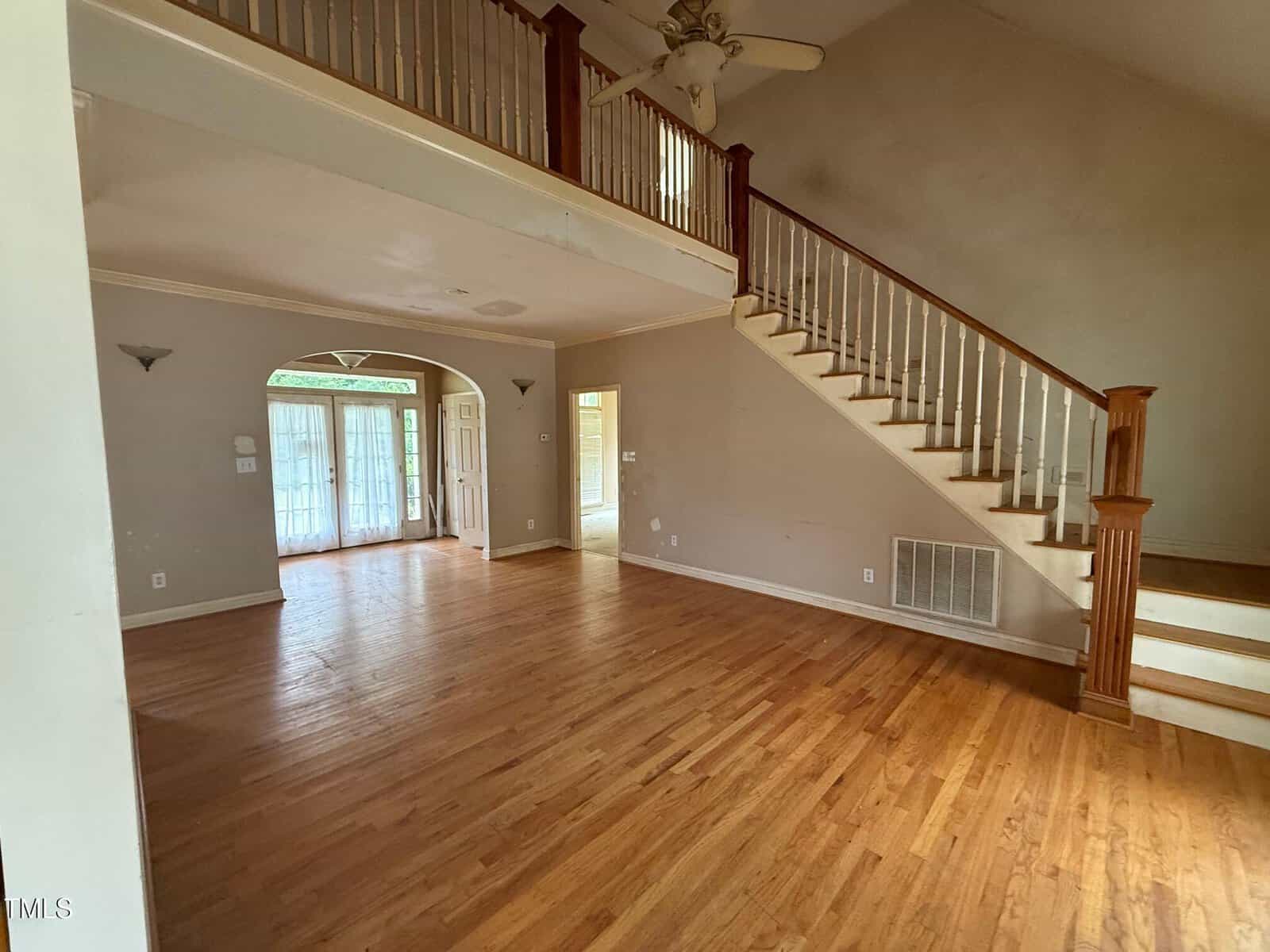 Spacious, empty living room with hardwood floors, high ceiling, a staircase with wooden railing, and an upper-level balcony. Sunlight enters through glass doors at the far end of the room.