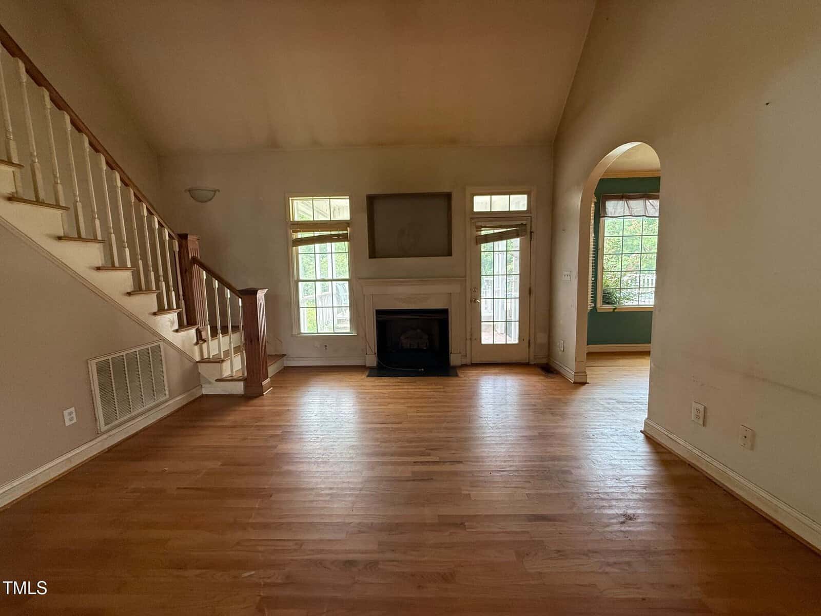 A mostly empty living room with hardwood floors, a fireplace centered on the far wall, a staircase on the left, and large windows letting in natural light. An open archway leads to another room on the right.