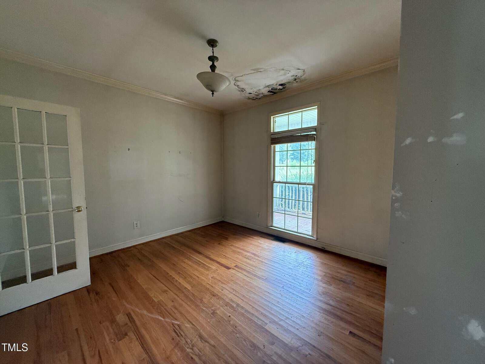 Empty room with hardwood floor, white walls, a ceiling light, and a glass-paned door. There is water damage on the ceiling above a window with a door leading outside. Light enters from the window.