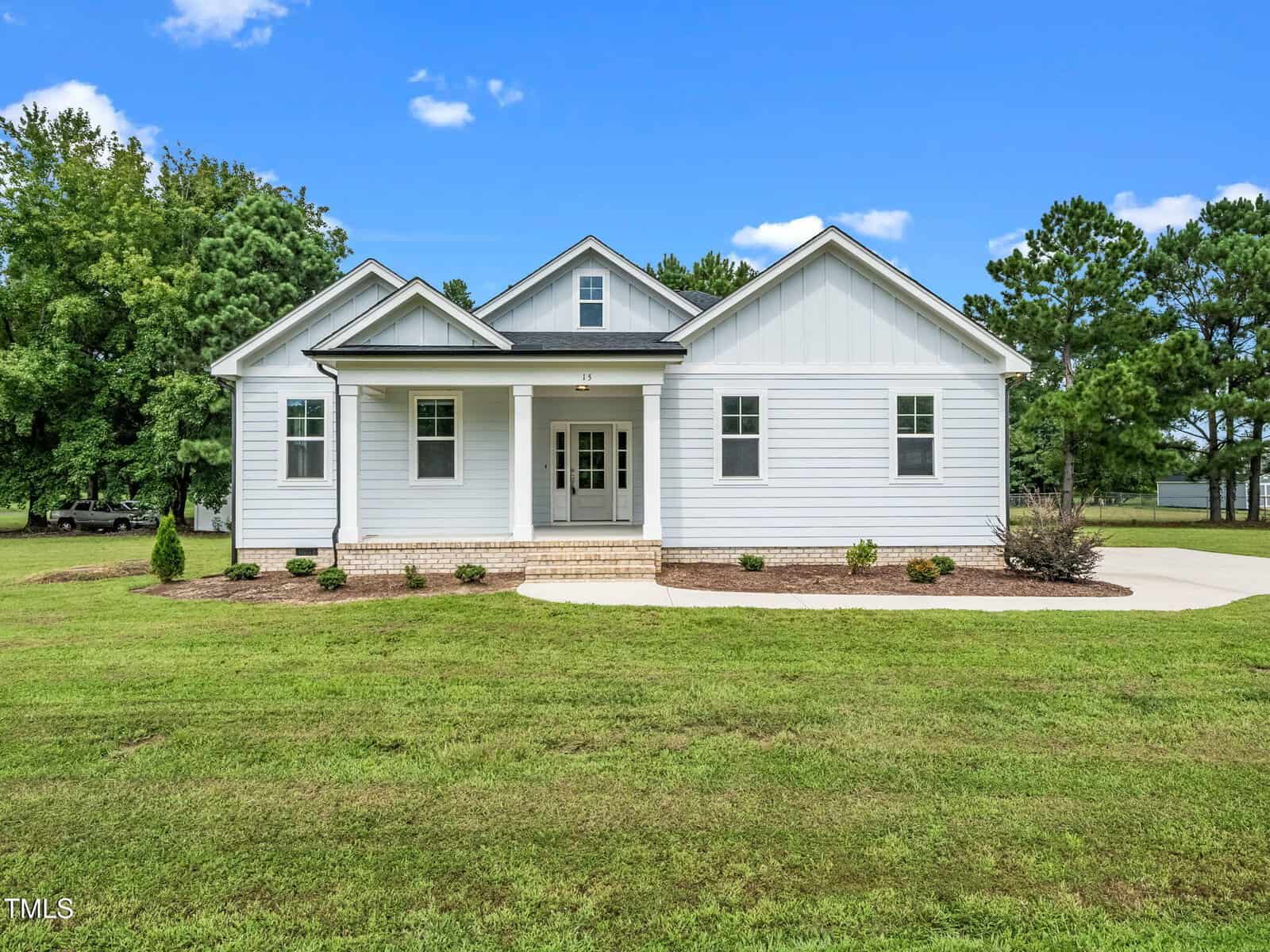 A single-story white house with a gray roof sits on a large grassy lawn at 15 Bella Lane, Zebulon. The house features a covered front porch, three front-facing gables, and a curved concrete walkway. Trees are visible in the background under a blue sky.