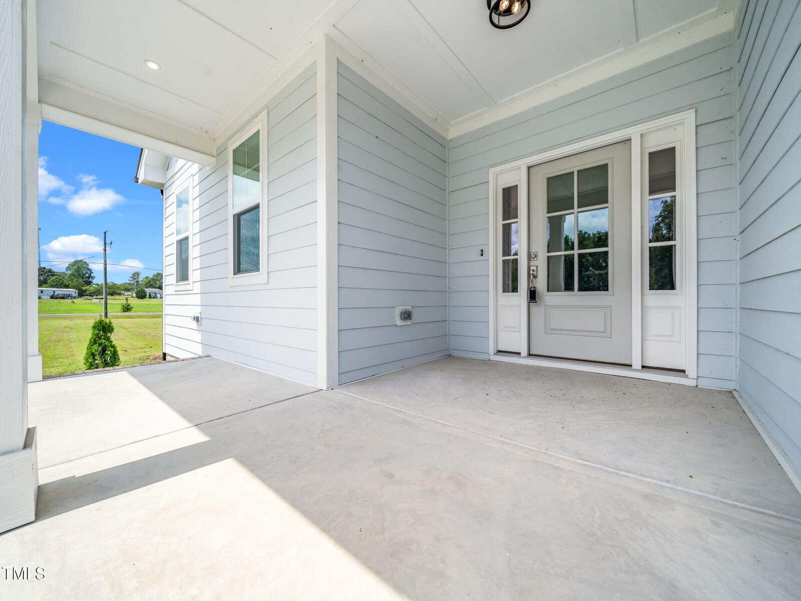 Covered front porch with light gray siding, white trim, and a white front door with glass panels. Concrete floor extends to the edge, and bright daylight highlights the clean, open entry space.