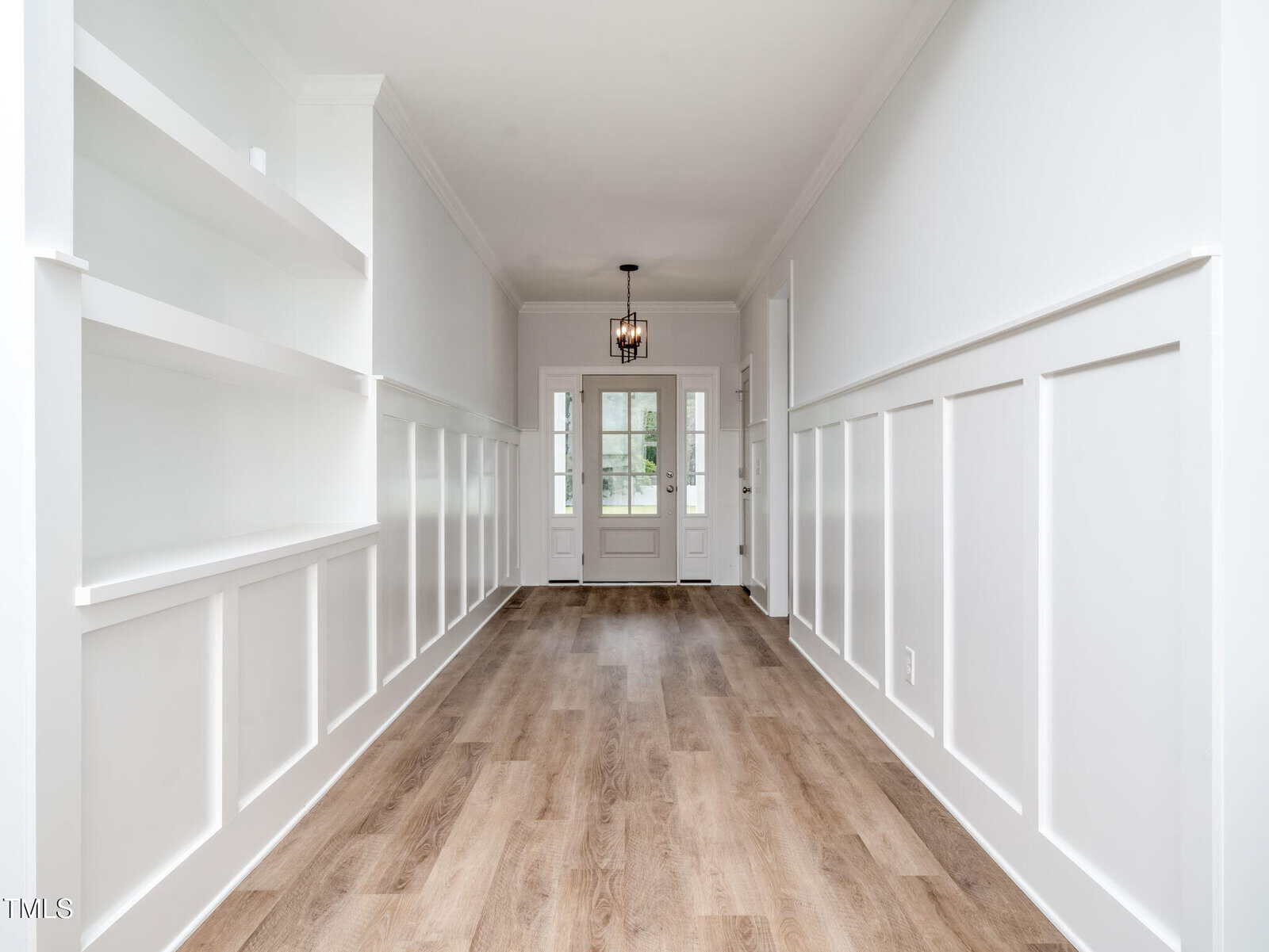 A bright hallway with white paneled walls, built-in shelves on the left, light wood flooring, and a modern pendant light. Double doors with glass panes are at the far end, letting in natural light.
