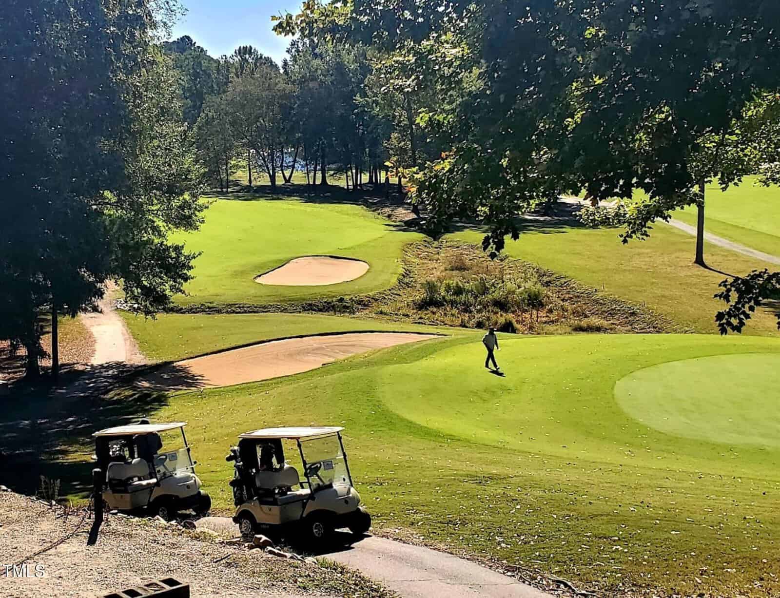 A golf course with lush green fairways, sand bunkers, and one person walking on the grass. Two golf carts are parked on a paved path in the foreground, surrounded by trees and sunlight.