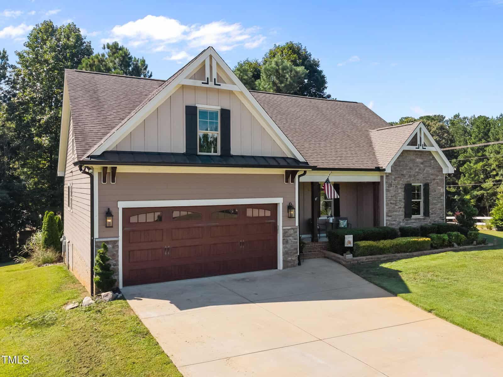 A single-story suburban house with beige siding, stone accents, a prominent gable, and a two-car garage with wooden-style doors. The front yard is landscaped with grass and small shrubs. An American flag hangs near the entryway.