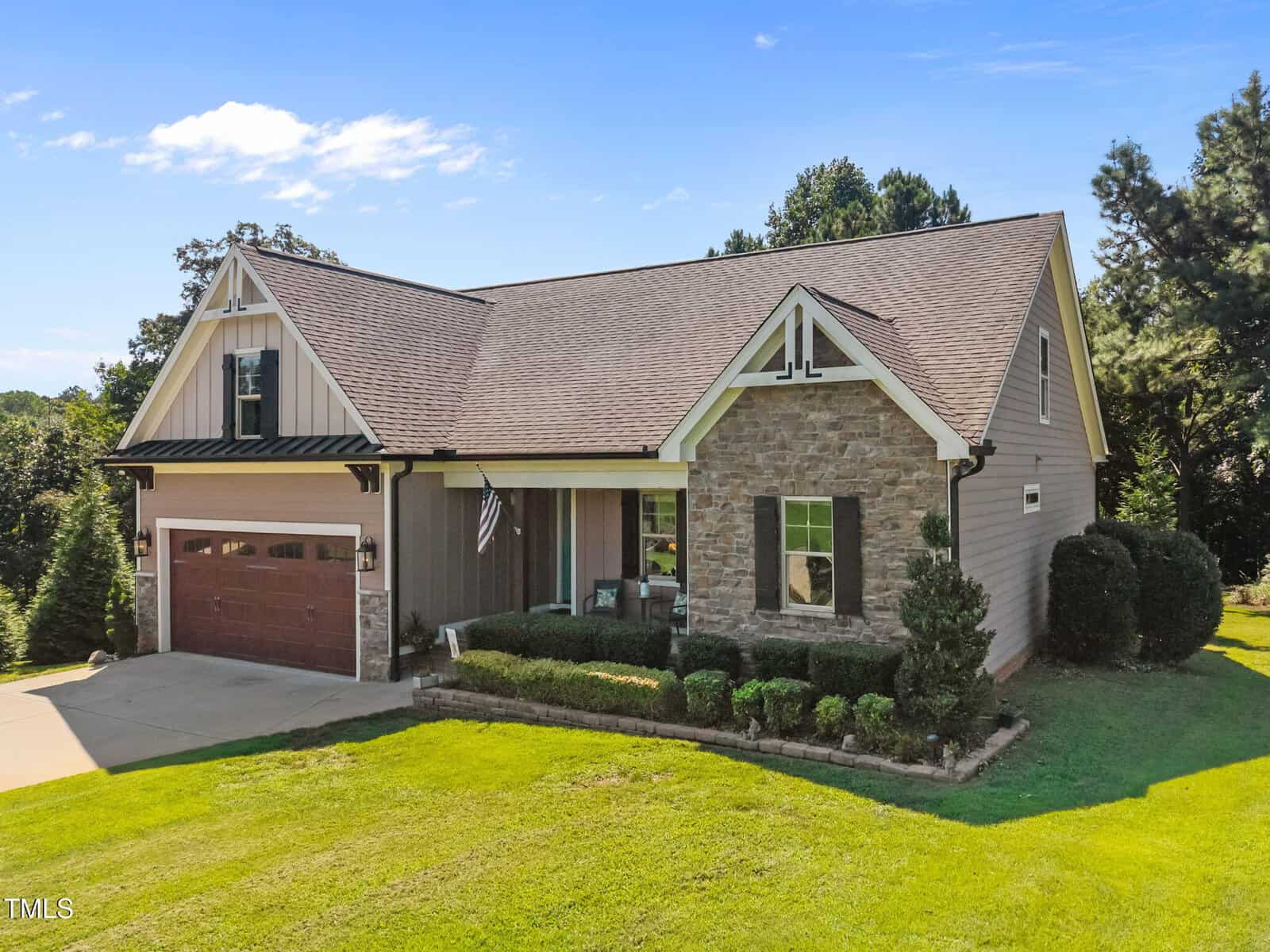 A single-story house with a double garage, stone and siding exterior, a gabled roof, and well-maintained landscaping, with a small front porch and a U.S. flag by the entrance.