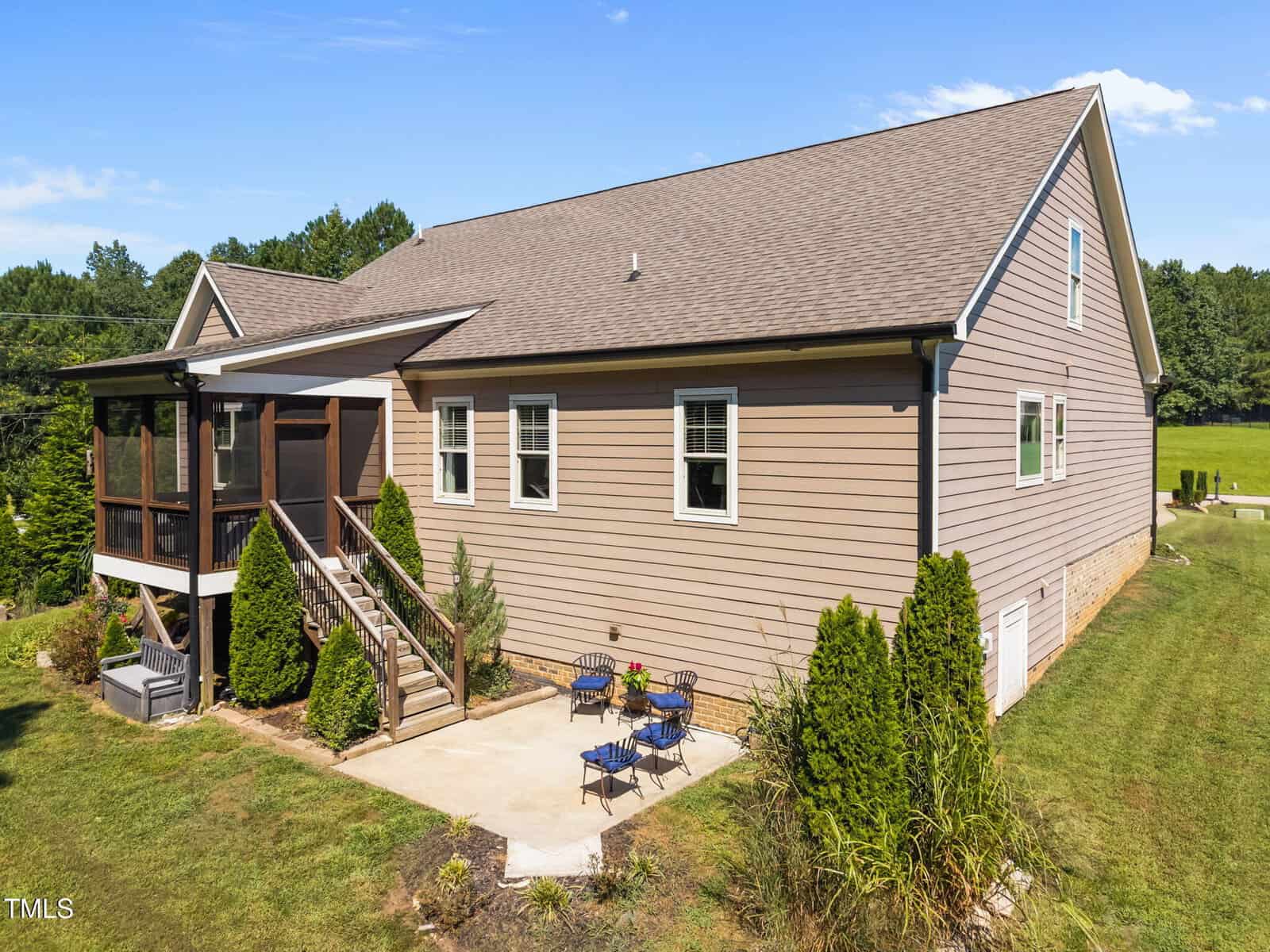 A beige single-story house with white trim, a screened-in porch with stairs, and a small concrete patio with blue chairs, set on a grassy lawn with shrubs and trees in the background.