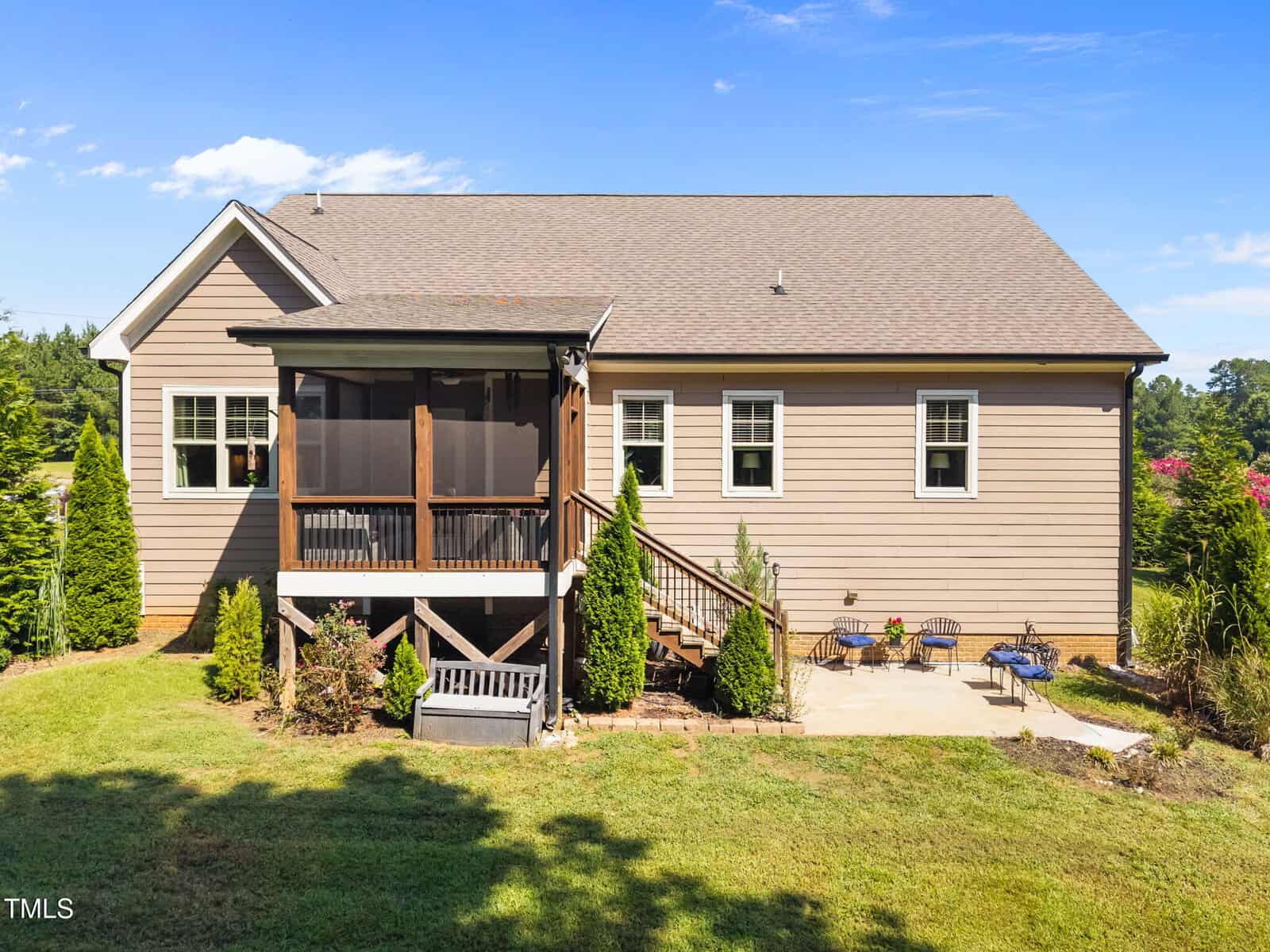 A single-story house with tan siding and a brown shingle roof. There is a screened porch with stairs, several windows, a small bench, and a seating area with two chairs and a table on a grassy lawn.