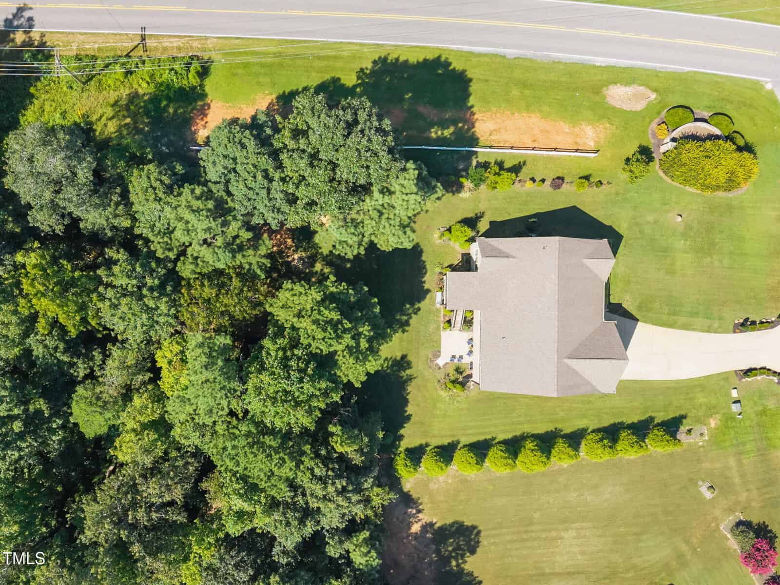 Aerial view of a single-story house with a gray roof, a curved driveway, and a large lawn. The property is bordered by dense trees on one side and a road at the top of the image.