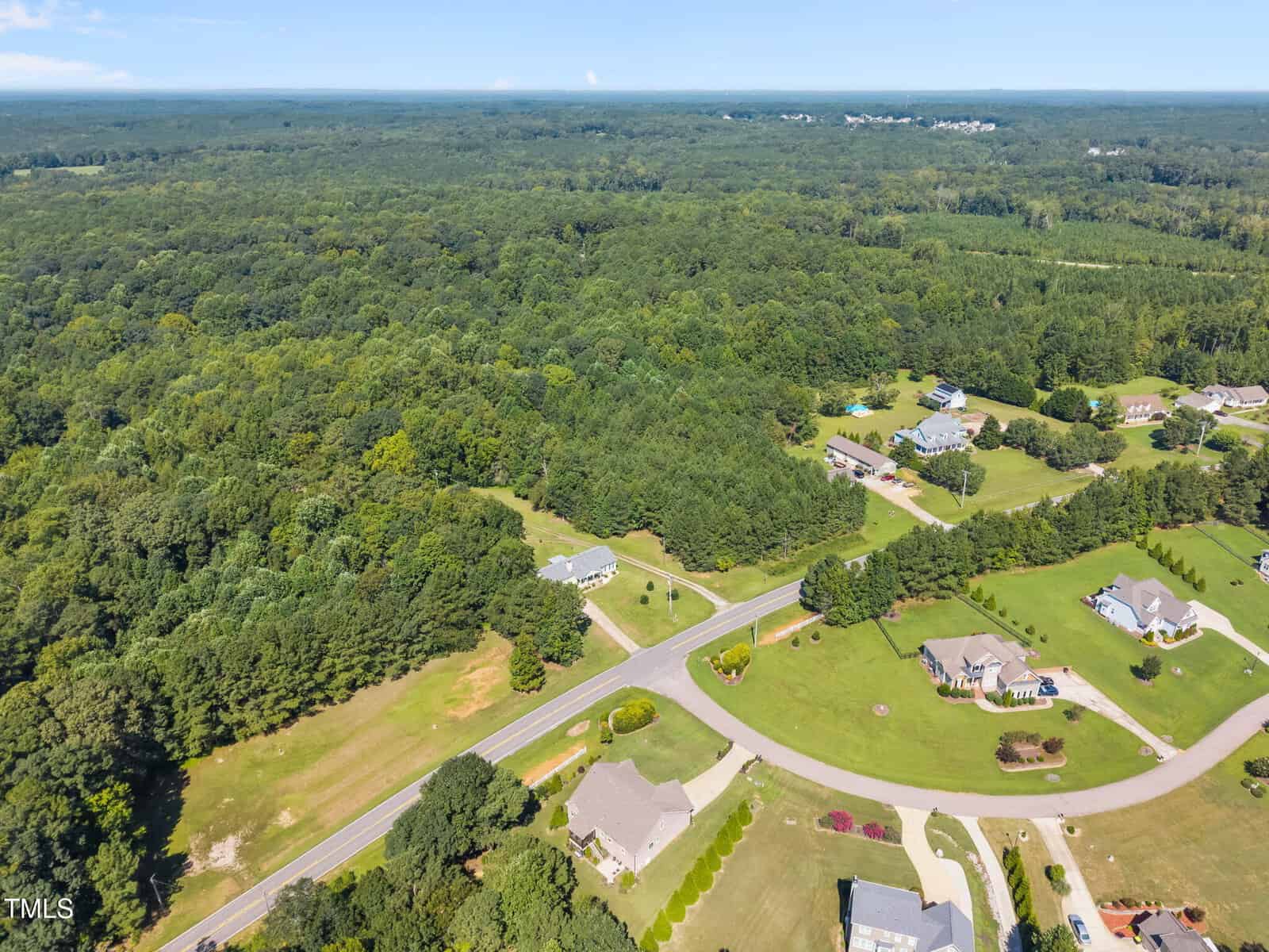 Aerial view of a residential neighborhood with houses, lawns, and curved roads next to a large, densely wooded forest under a clear blue sky.