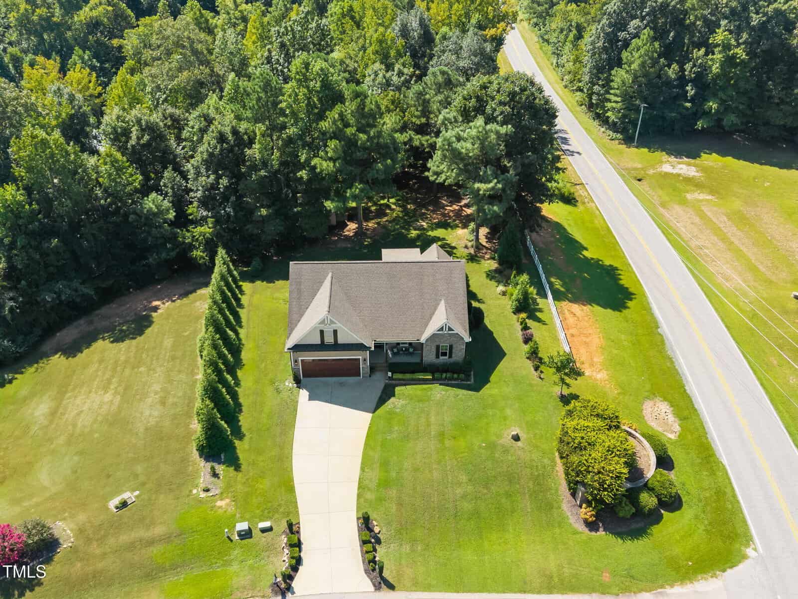Aerial view of a single-story house with a driveway, green lawn, trees surrounding the property, and a road running beside it on a sunny day.