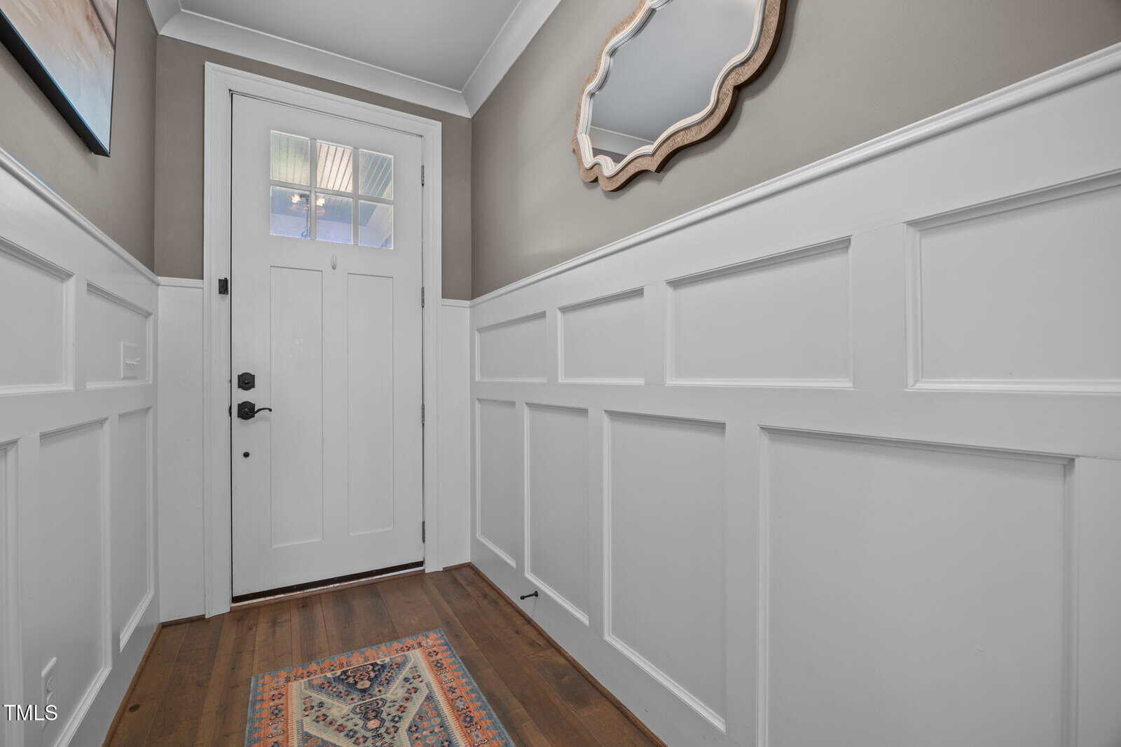 A hallway with white wainscoting, a wooden floor, a patterned rug, a white front door with window panes, taupe walls, and a decorative mirror hanging on the right wall.