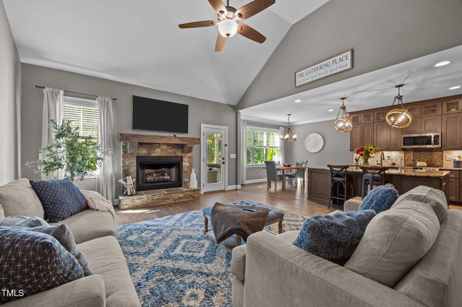 Bright living room with gray couches, blue accents, stone fireplace, wall-mounted TV, ceiling fan, and open layout to a kitchen with dark cabinets and dining area. Large windows let in natural light.