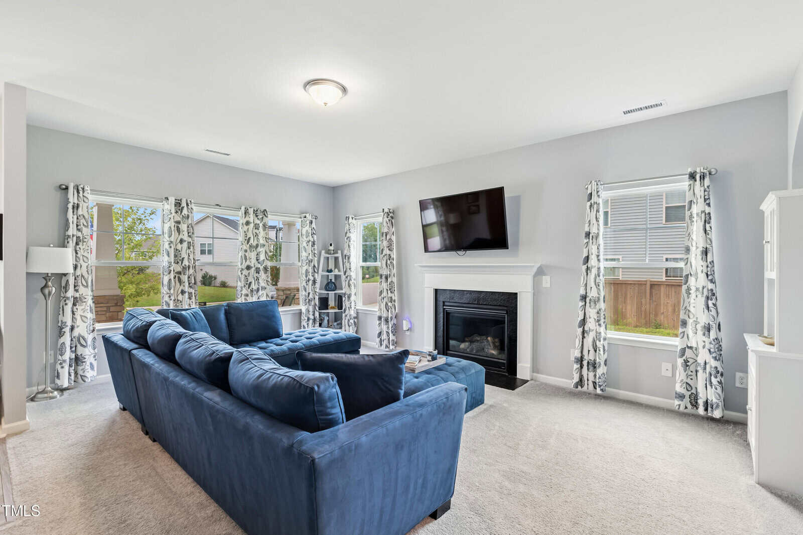 A living room with light gray walls, a blue sectional sofa, a wall-mounted TV above a fireplace, several windows with patterned curtains, and beige carpet flooring. A backyard and neighboring house are visible through the windows.