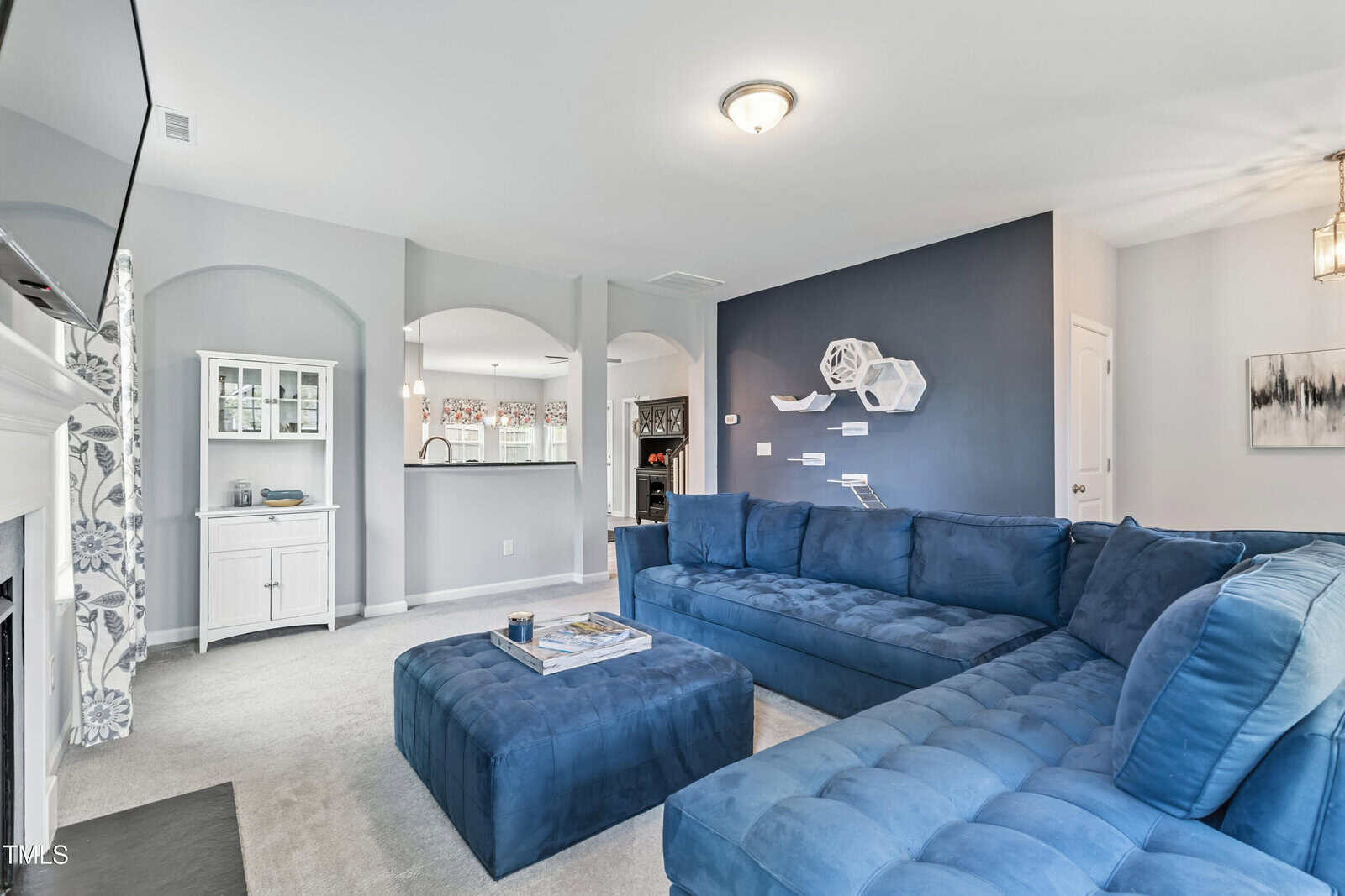 A living room with a blue sectional sofa, matching ottoman, dark accent wall, hexagonal shelves, and an open view into the kitchen with arched pass-throughs. Light gray walls and carpet complete the space.