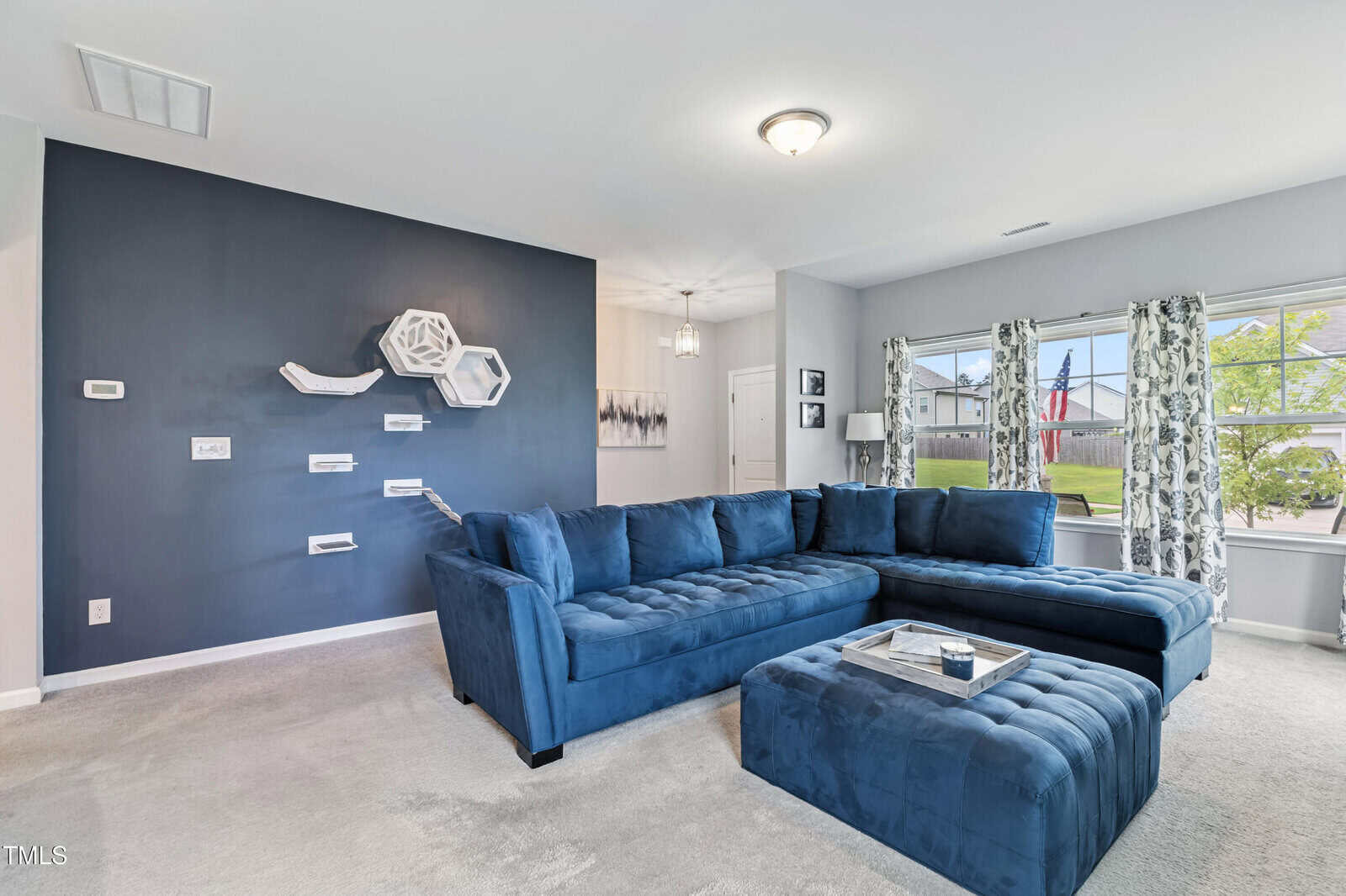 A modern living room with a large blue sectional sofa, matching ottoman, tray, and a blue accent wall with geometric shelves. Windows with patterned curtains let in natural light. Neutral walls and carpet complete the space.