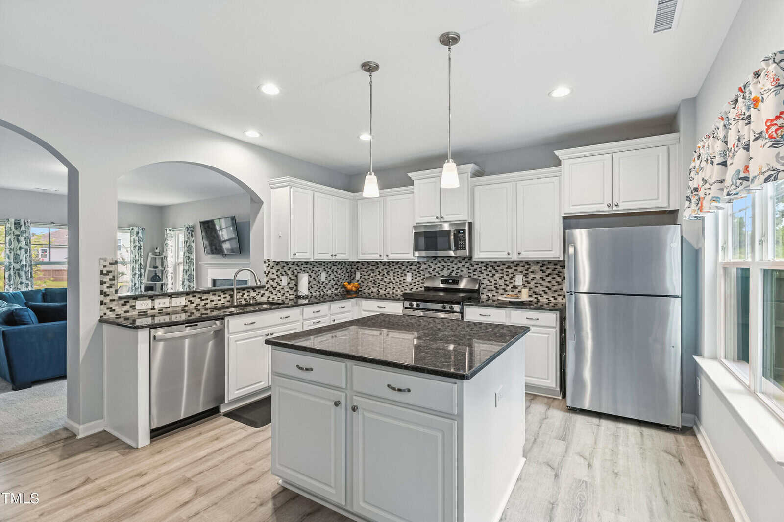 Modern kitchen with white cabinets, stainless steel appliances, a gray and black mosaic tile backsplash, center island with dark countertop, pendant lights, and a window with floral curtains. Light wood flooring throughout.