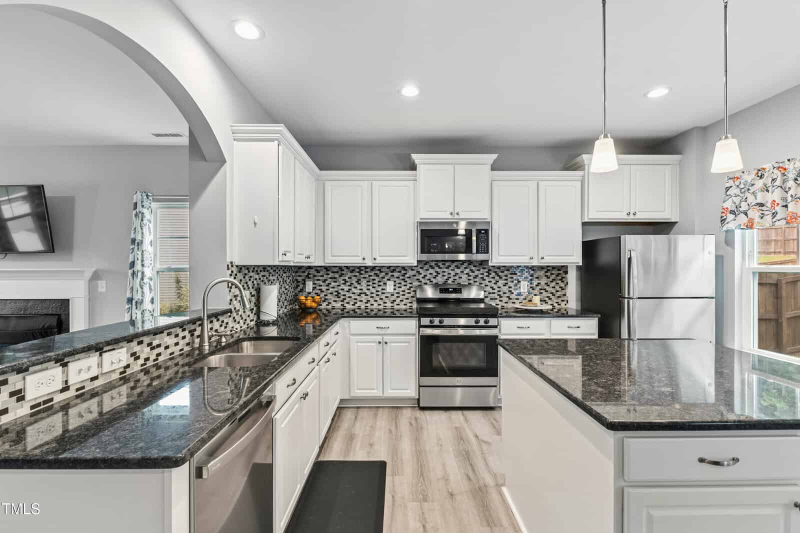 Modern kitchen with white cabinets, stainless steel appliances, black granite countertops, mosaic tile backsplash, pendant lighting, and wood flooring. An arched opening reveals a living room with a fireplace and wall-mounted TV.