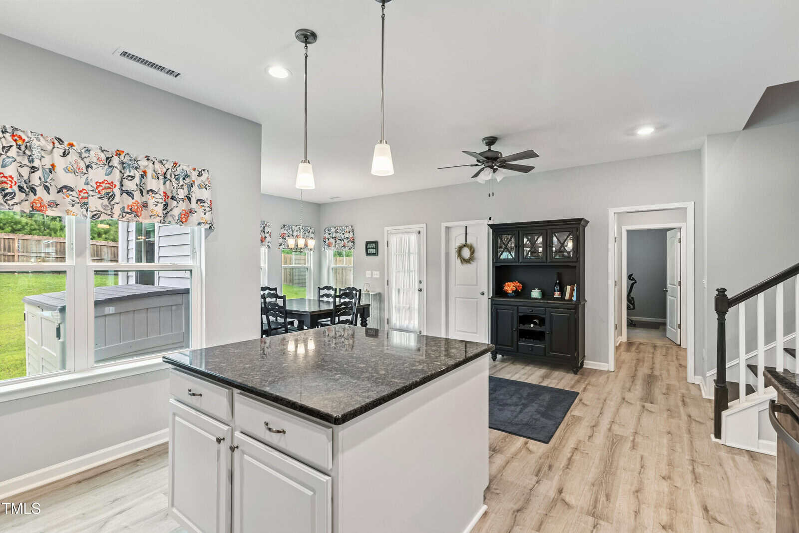 Bright kitchen with a large granite island, white cabinets, wood floors, a black dining hutch, dining table by windows with colorful valances, ceiling fan, and a view of a fenced backyard through multiple windows.