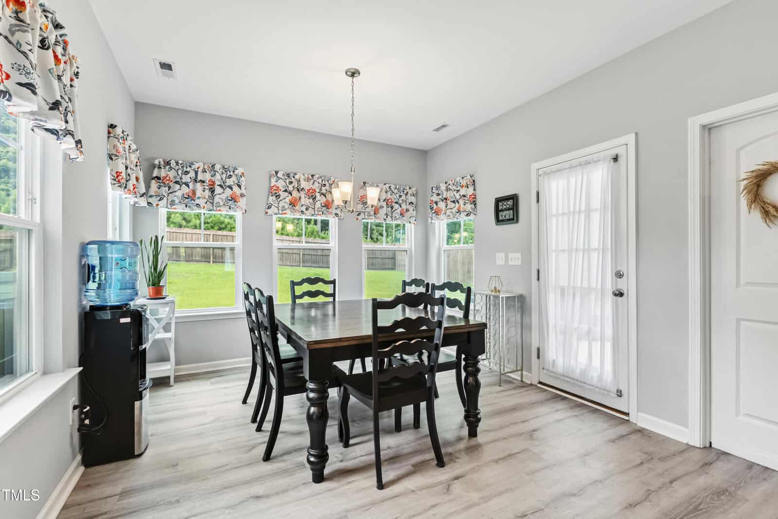 A bright dining room with light gray walls, wood flooring, a dark wooden table with six black chairs, floral curtains, a water dispenser, and large windows overlooking a green yard. A glass door leads outside.