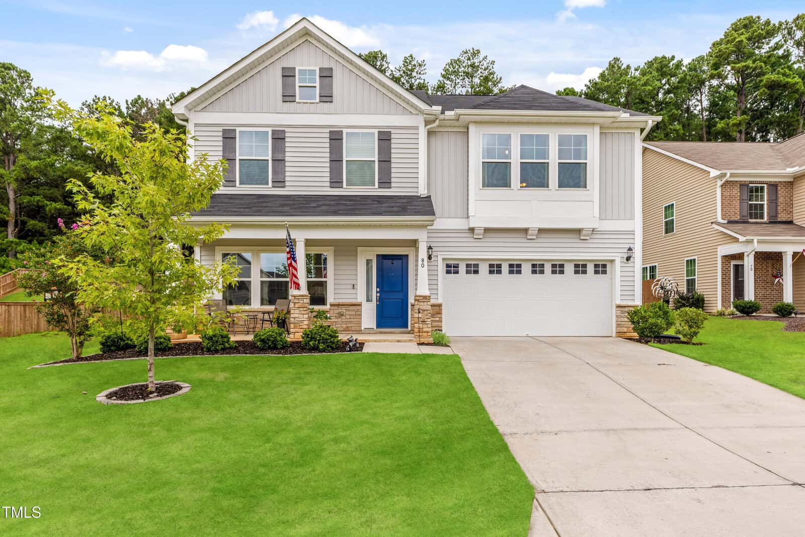 Two-story suburban house with white siding, gray shutters, and a blue front door. It has a double garage, a small covered porch with an American flag, manicured lawn, and young trees, with neighboring houses nearby.