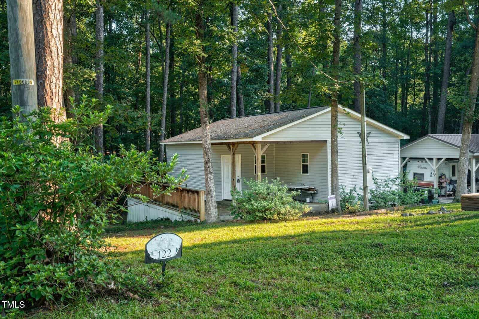 A small white house with a covered porch sits among tall trees and greenery. A wooden fence runs alongside it, and a sign with the number 122 is on the grassy lawn. Sunlight filters through the forested background.