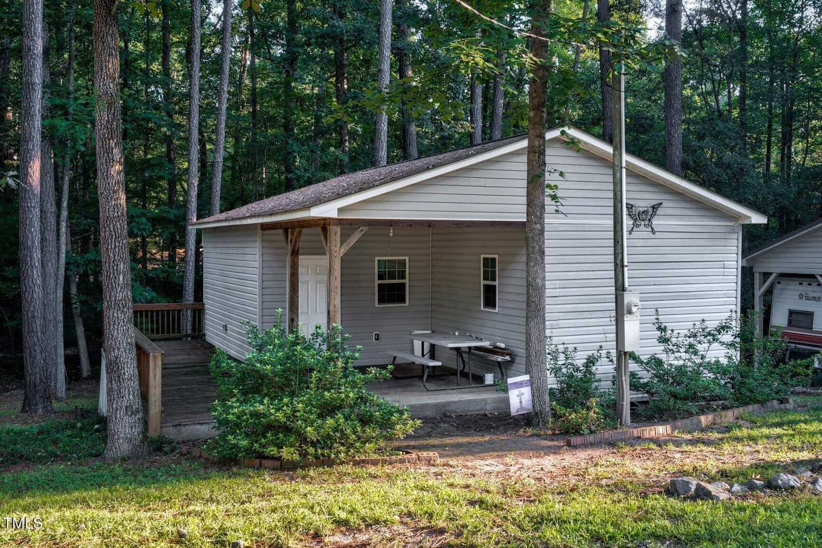 A small, white, single-story house with a covered front porch, picnic table, and ramp, surrounded by tall trees and greenery in a wooded area. Sunlight filters through the trees onto the grassy yard.