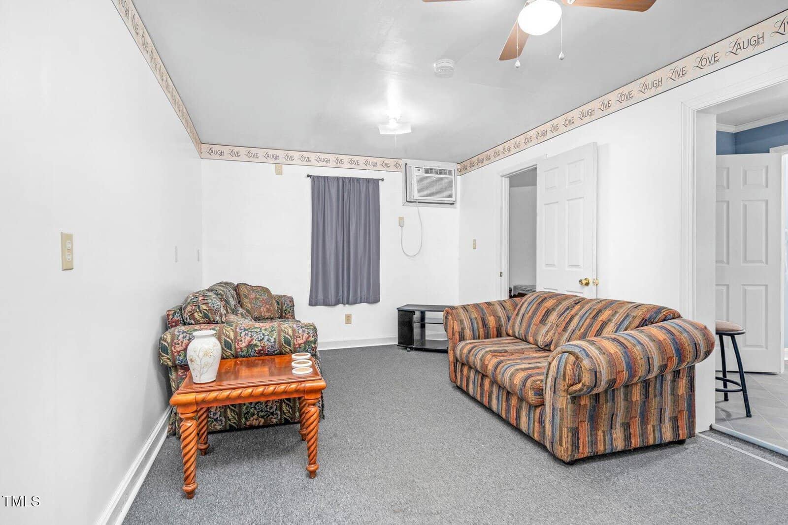 A small living room with two patterned sofas, a wooden coffee table, a window with a gray curtain, a ceiling fan, and a wall air conditioner. The walls are white, and there is writing along the border near the ceiling.