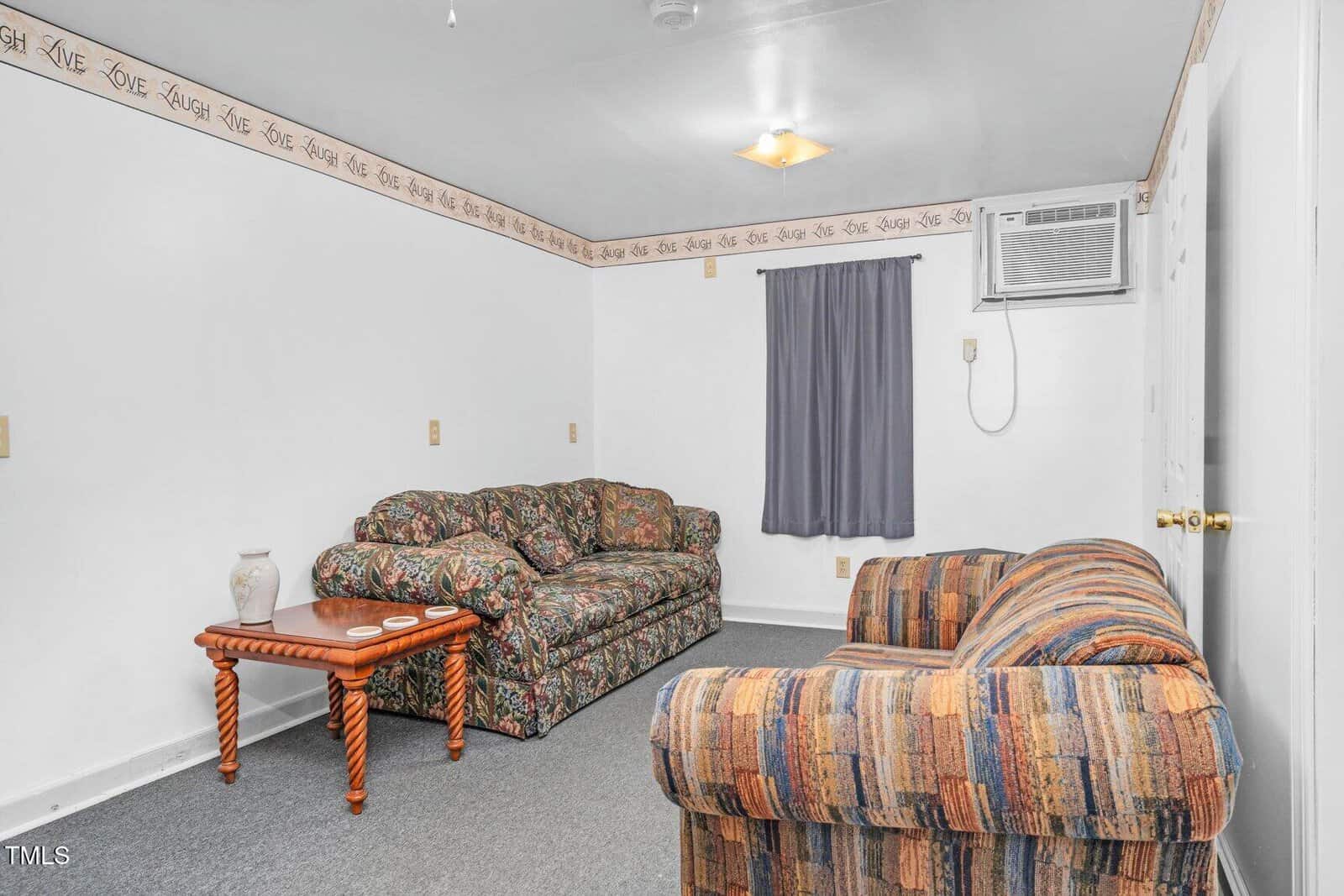 Small living room with patterned sofas, a wooden side table with two vases, gray carpet, white walls with a decorative border, a curtained window, ceiling light, and a wall-mounted air conditioning unit.