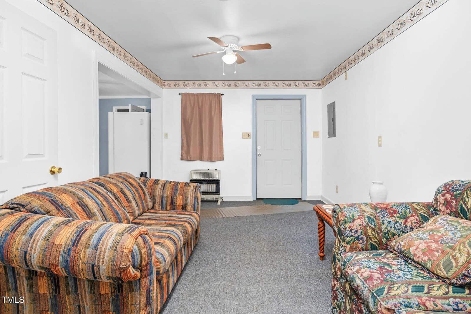 A living room with a patterned sofa and armchair, gray carpet, white walls, a ceiling fan, and a window with a brown curtain. The entry door is light blue and there is floral wallpaper trim along the ceiling.