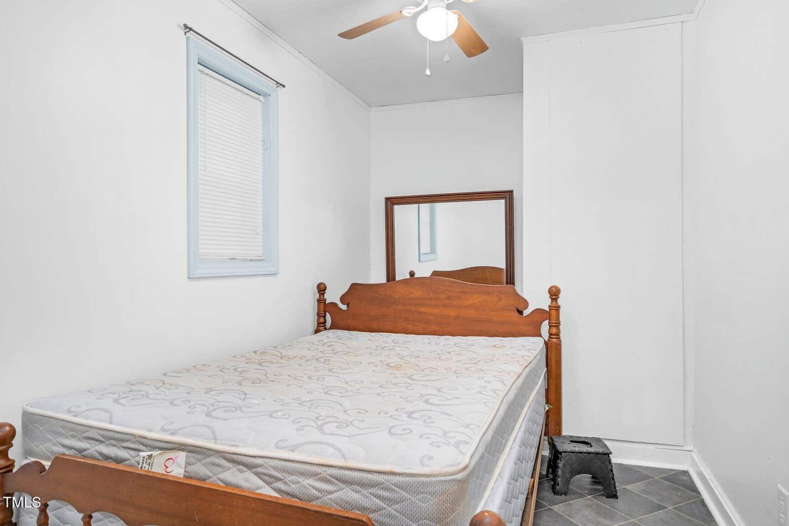 A small bedroom with a wooden bed and mattress, a large mirror against the back wall, a ceiling fan, a window with closed blinds, and a black step stool on a tiled floor. Walls are painted white.