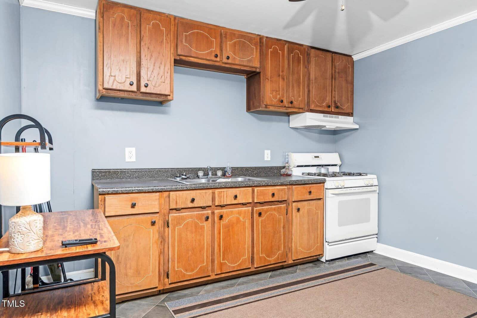 A small kitchen with wooden cabinets, a countertop with a sink, a white gas stove with range hood, and a brown rug. A wooden table with a lamp and phone is on the left side against blue walls.